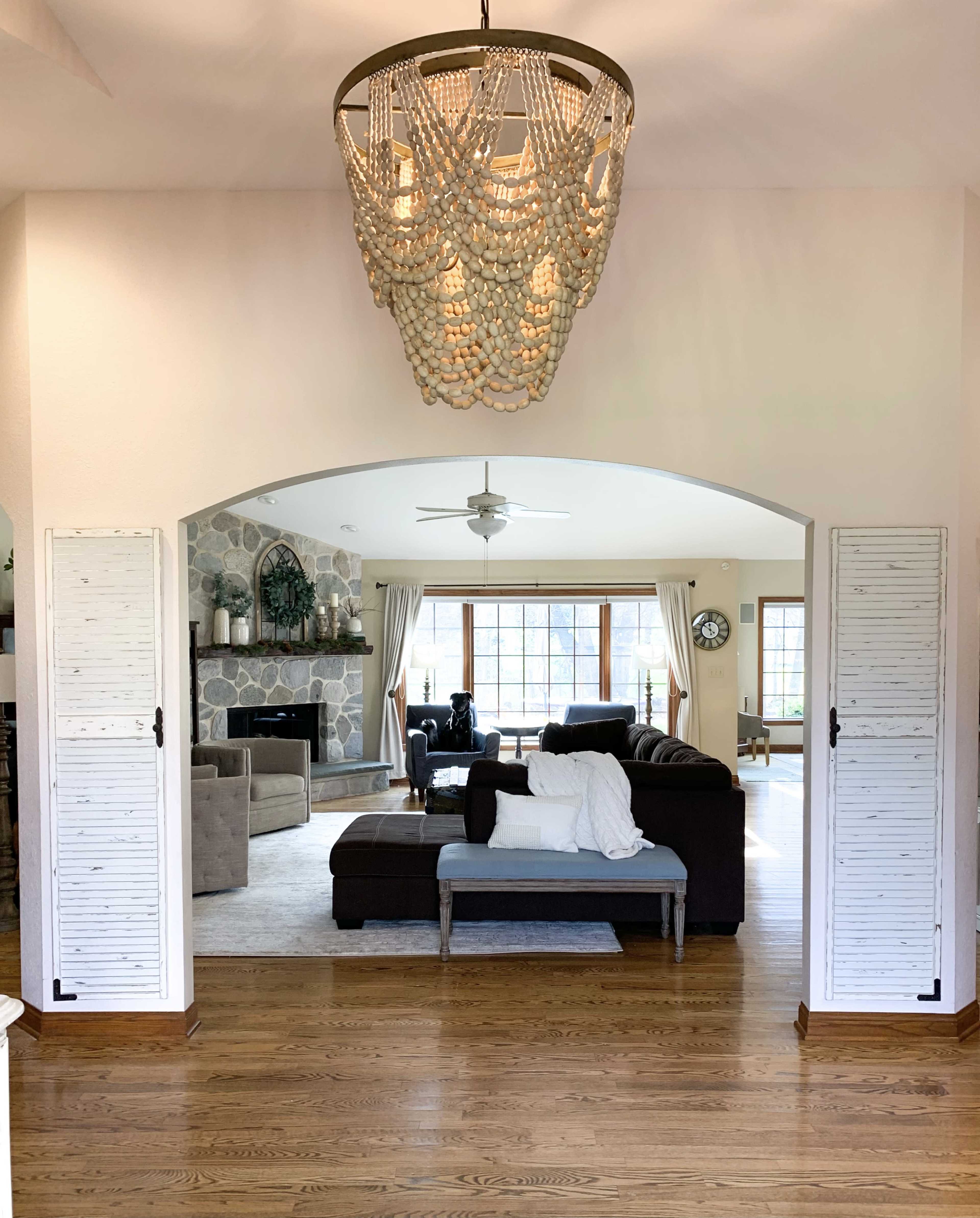 A spacious living room with a stone fireplace and a chandelier above, seen through an archway flanked by shuttered doors.