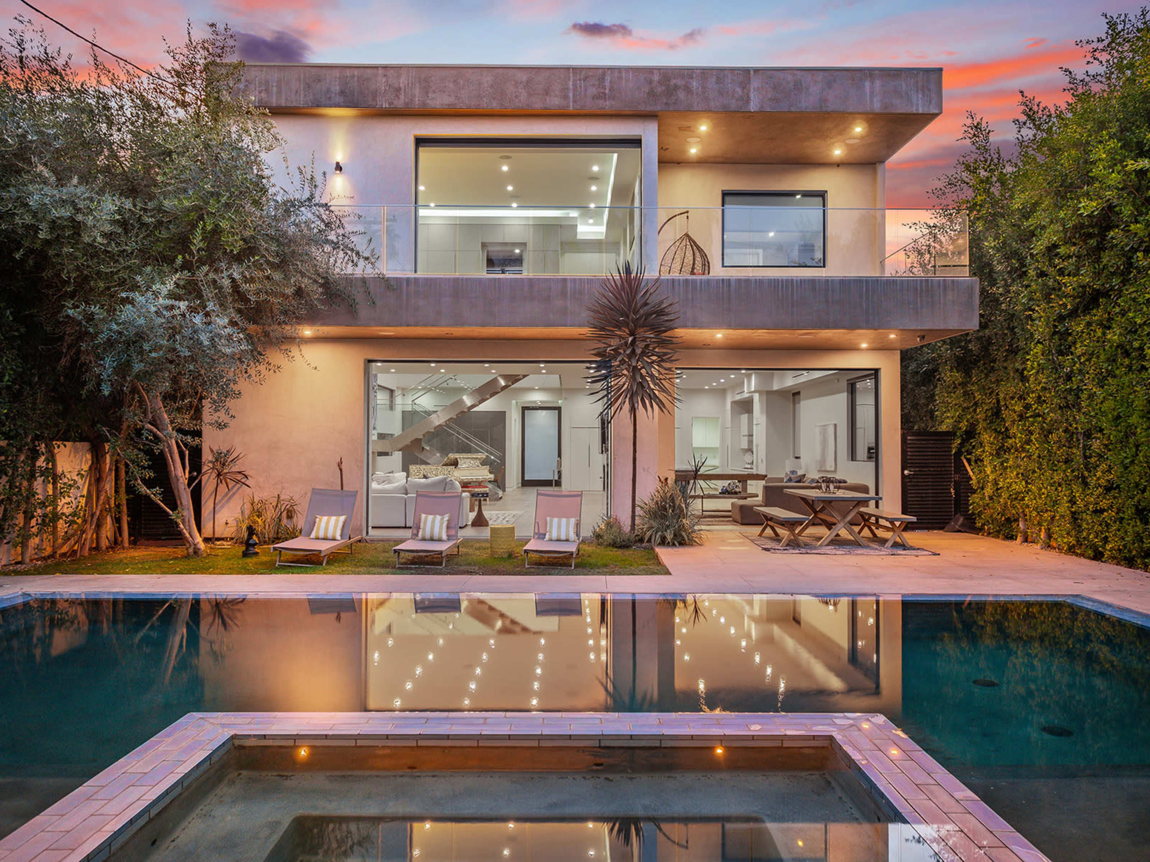 A modern two-story house with large windows is reflected in a swimming pool, surrounded by greenery and lounge chairs.