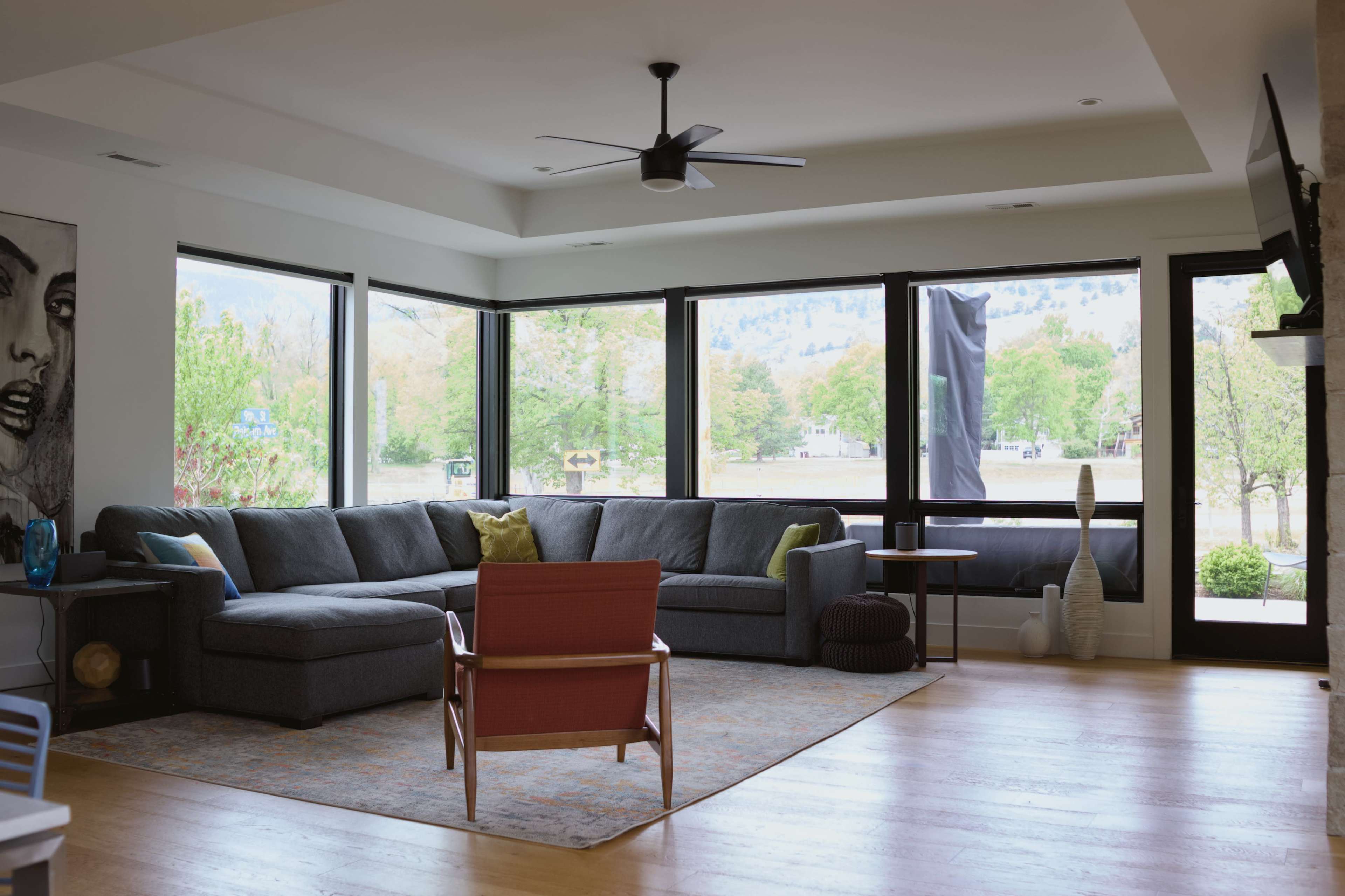 The image shows a modern living room with a large gray sectional sofa, a wooden chair, and big windows overlooking a green outdoor area.