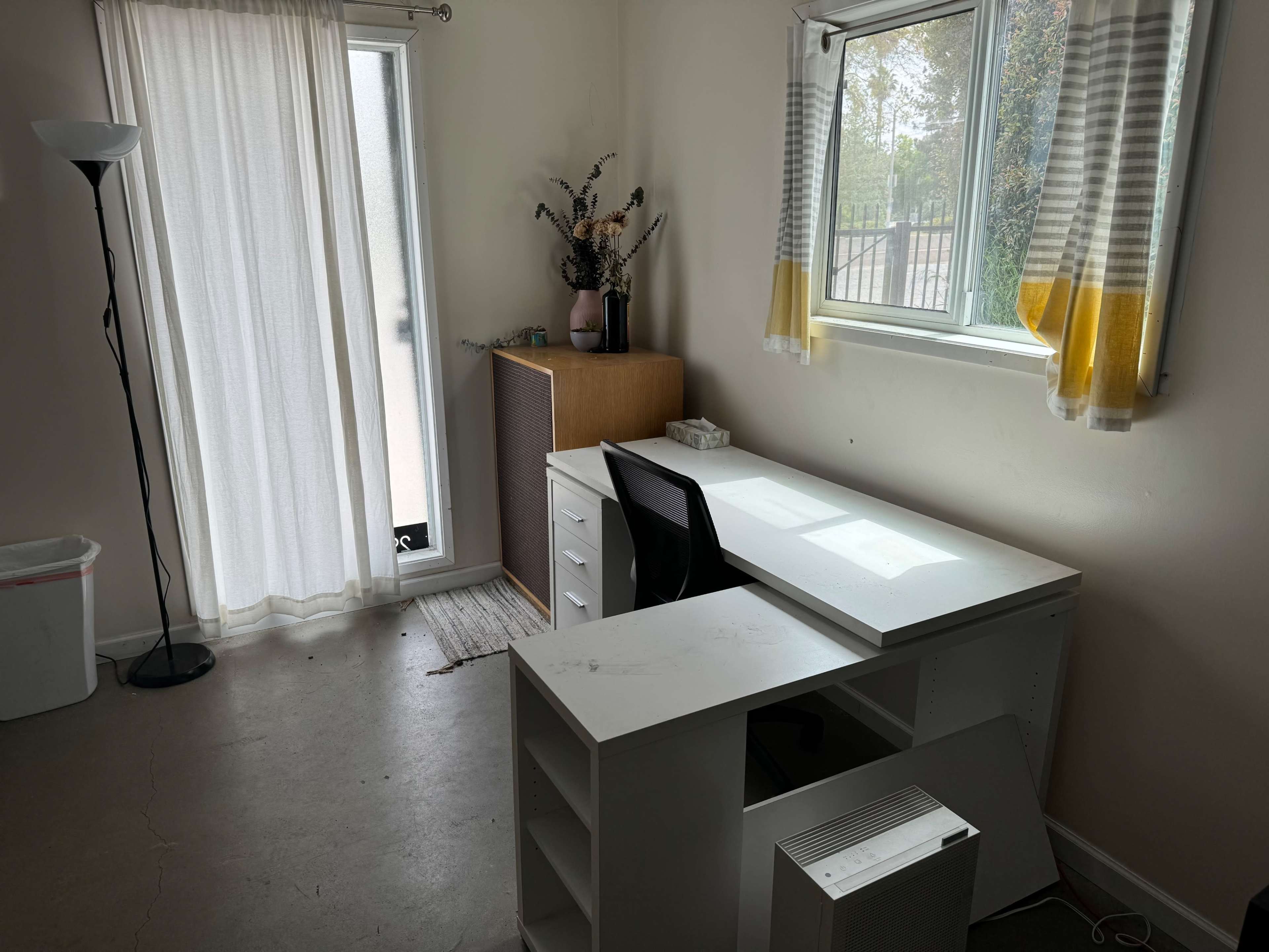 A simple office space with a white desk, a black chair, a cabinet, a lamp, and a window with sheer curtains.