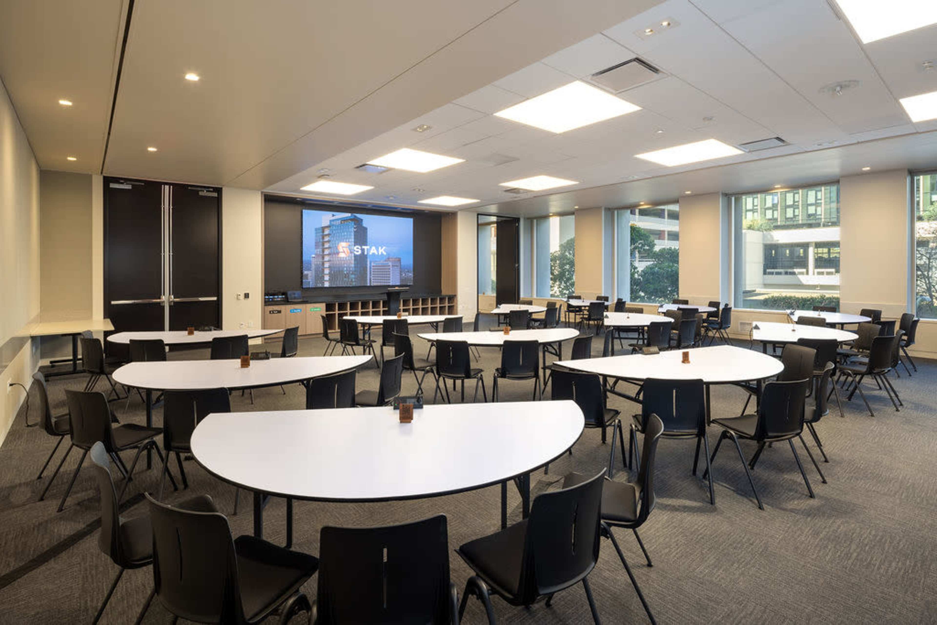 The image shows a modern conference room with several round tables arranged in a spacious layout, featuring a large screen displaying the word "STAK" on one wall.