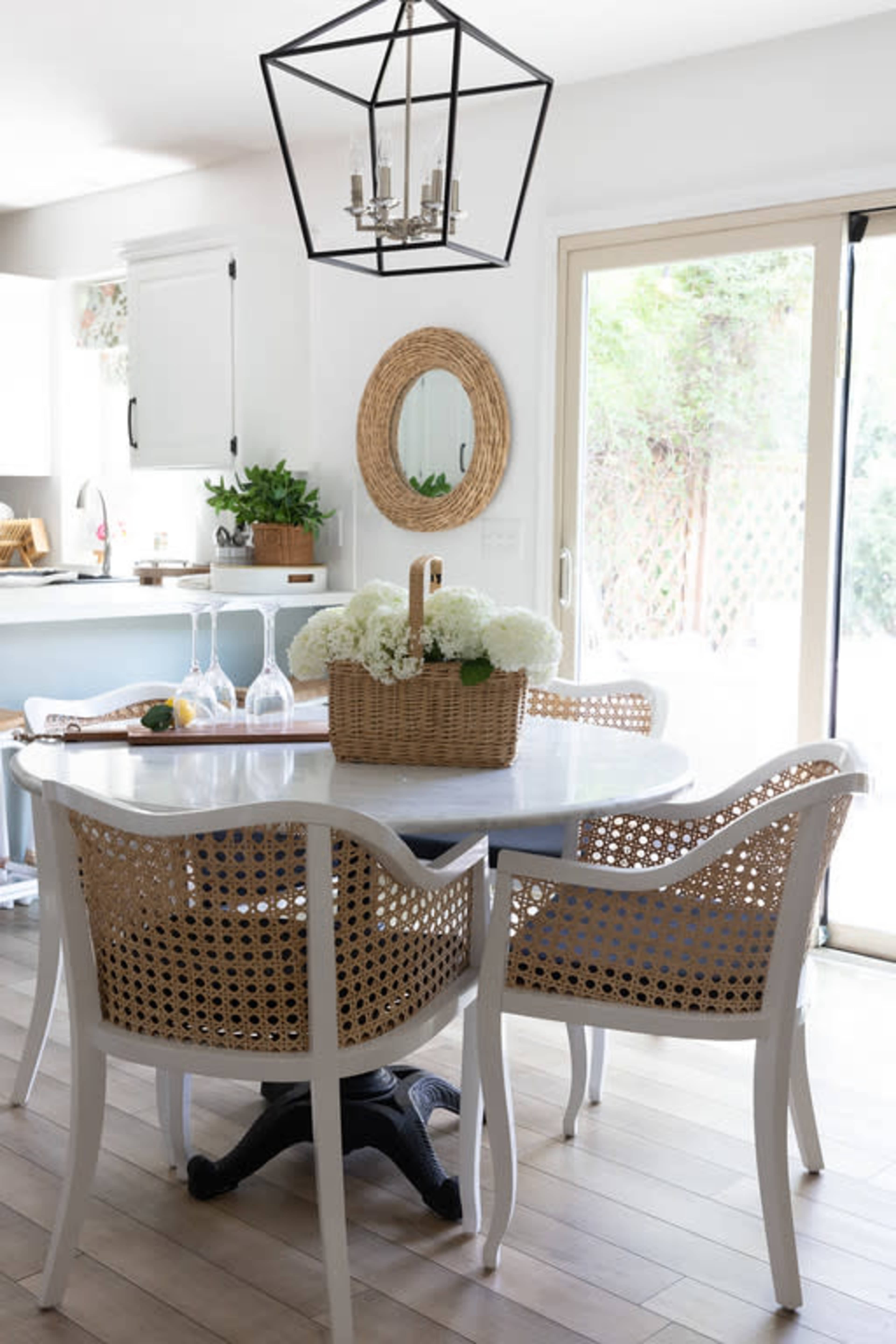 The image shows a bright dining area with a round white table surrounded by four rattan chairs, a woven basket of flowers as the centerpiece, and a sliding glass door leading to an outdoor space.