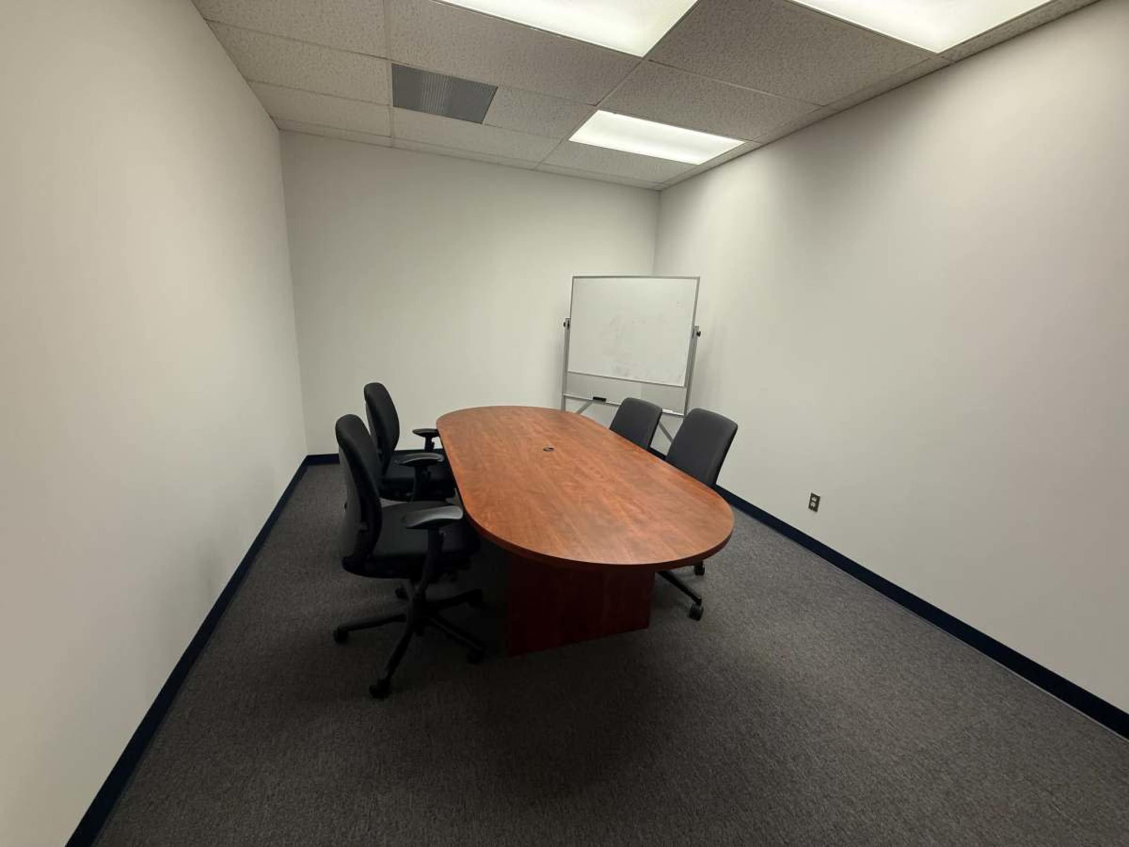 The image shows a small, empty conference room with a wooden table and four chairs surrounding it, along with a whiteboard on one wall.