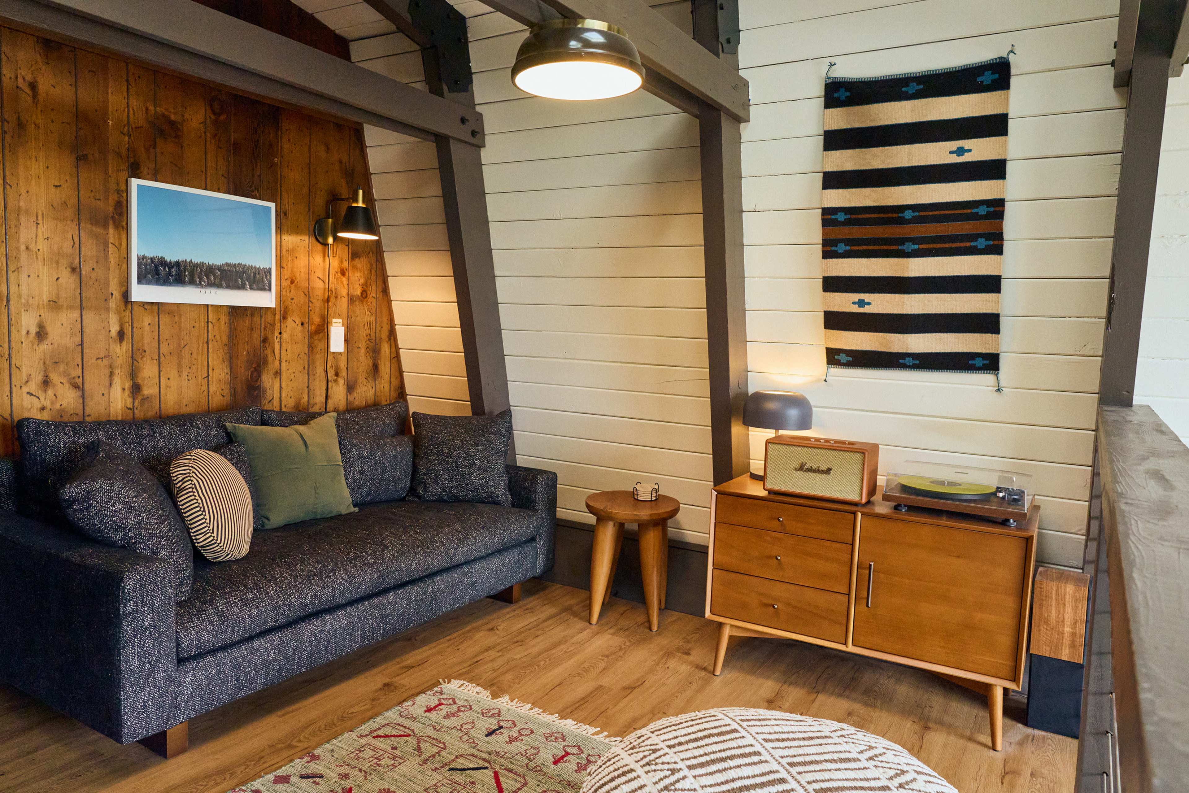 The image shows a cozy living room with a dark sofa, wooden accent wall, a small side table, and a record player on a mid-century modern console.