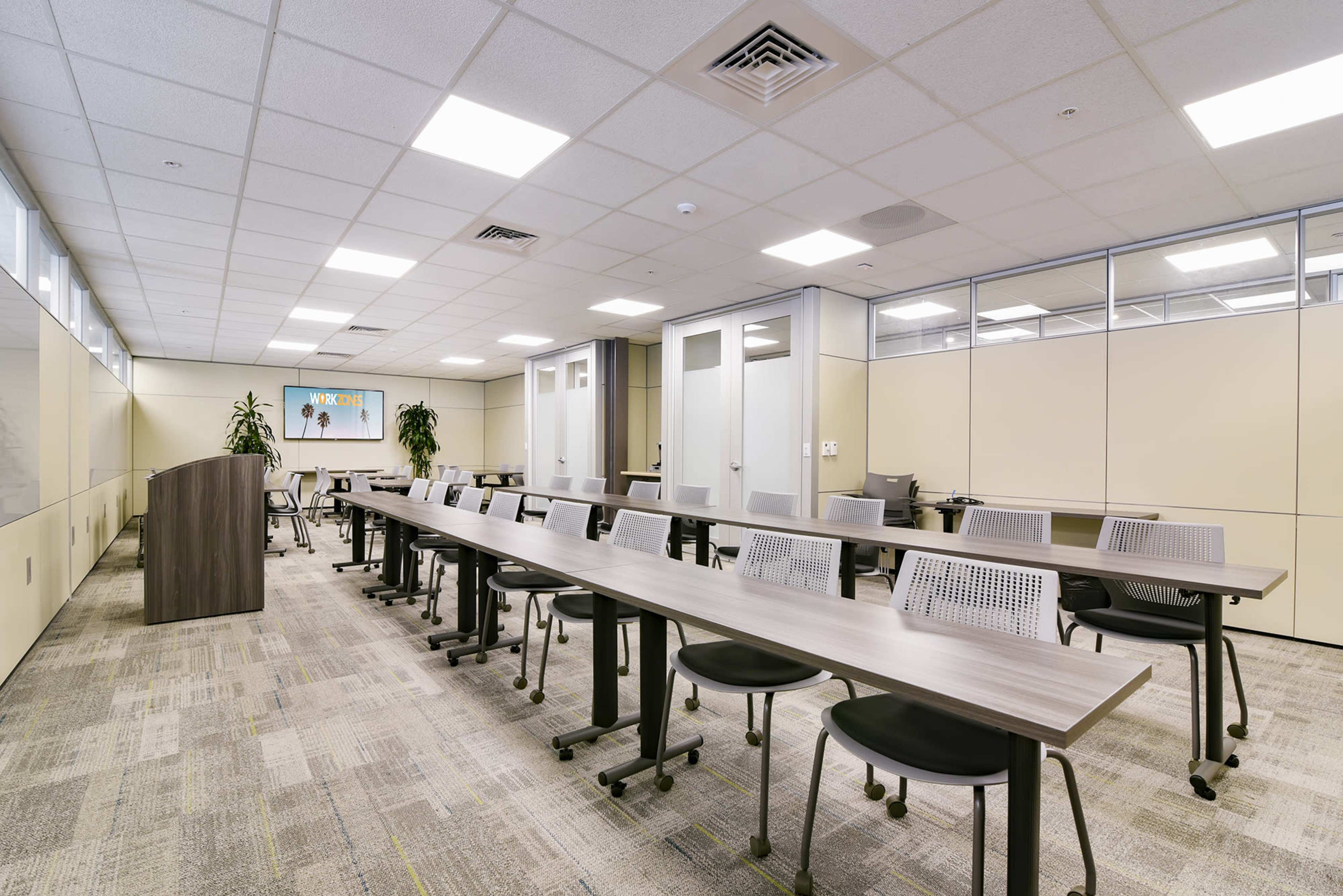 A modern conference room with long tables arranged in a row, accompanied by rolling chairs and plants in the corners.