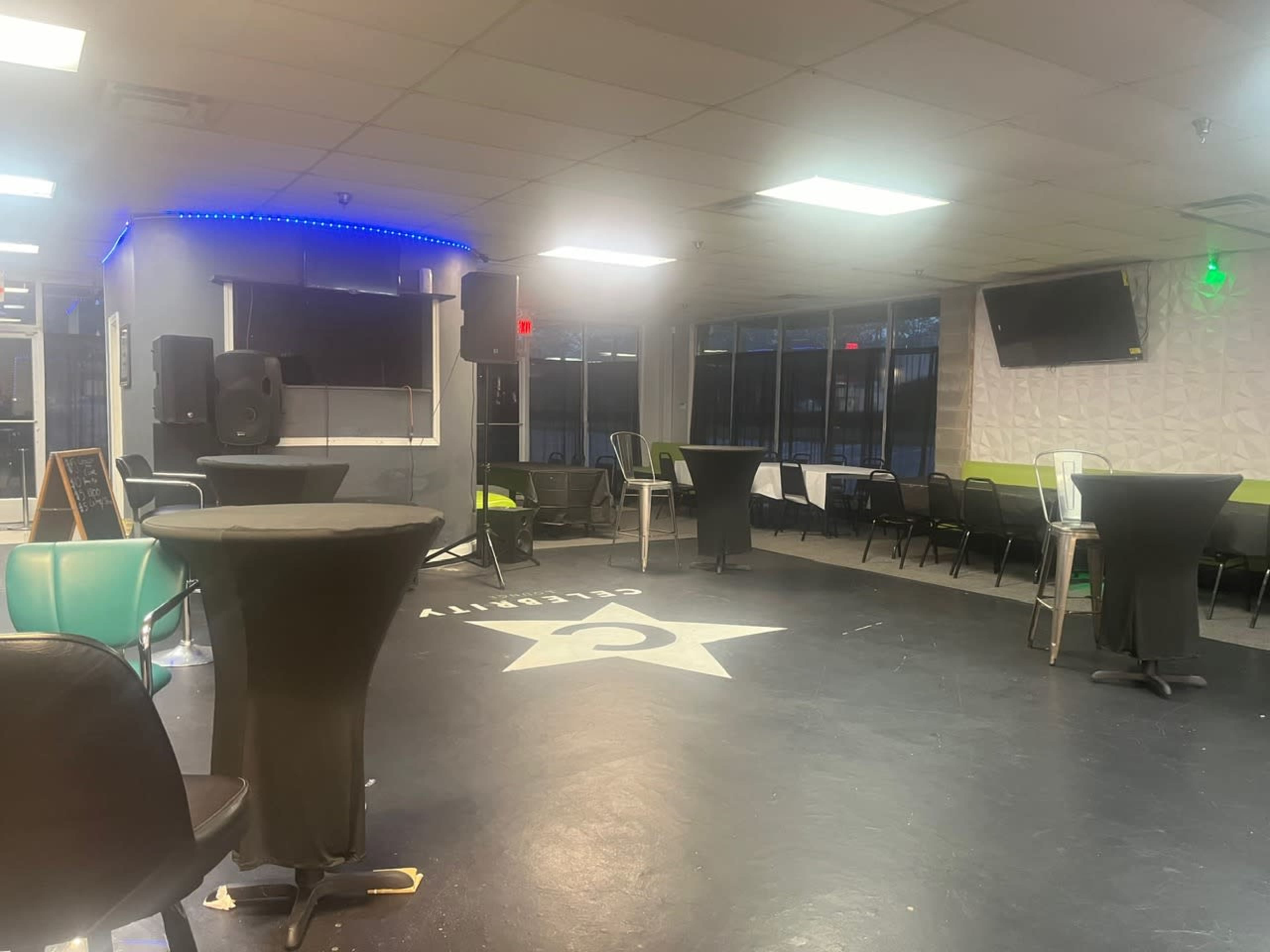 An empty event space with dark flooring, round tables covered with black cloth, and a stage setup with speakers and lighting.