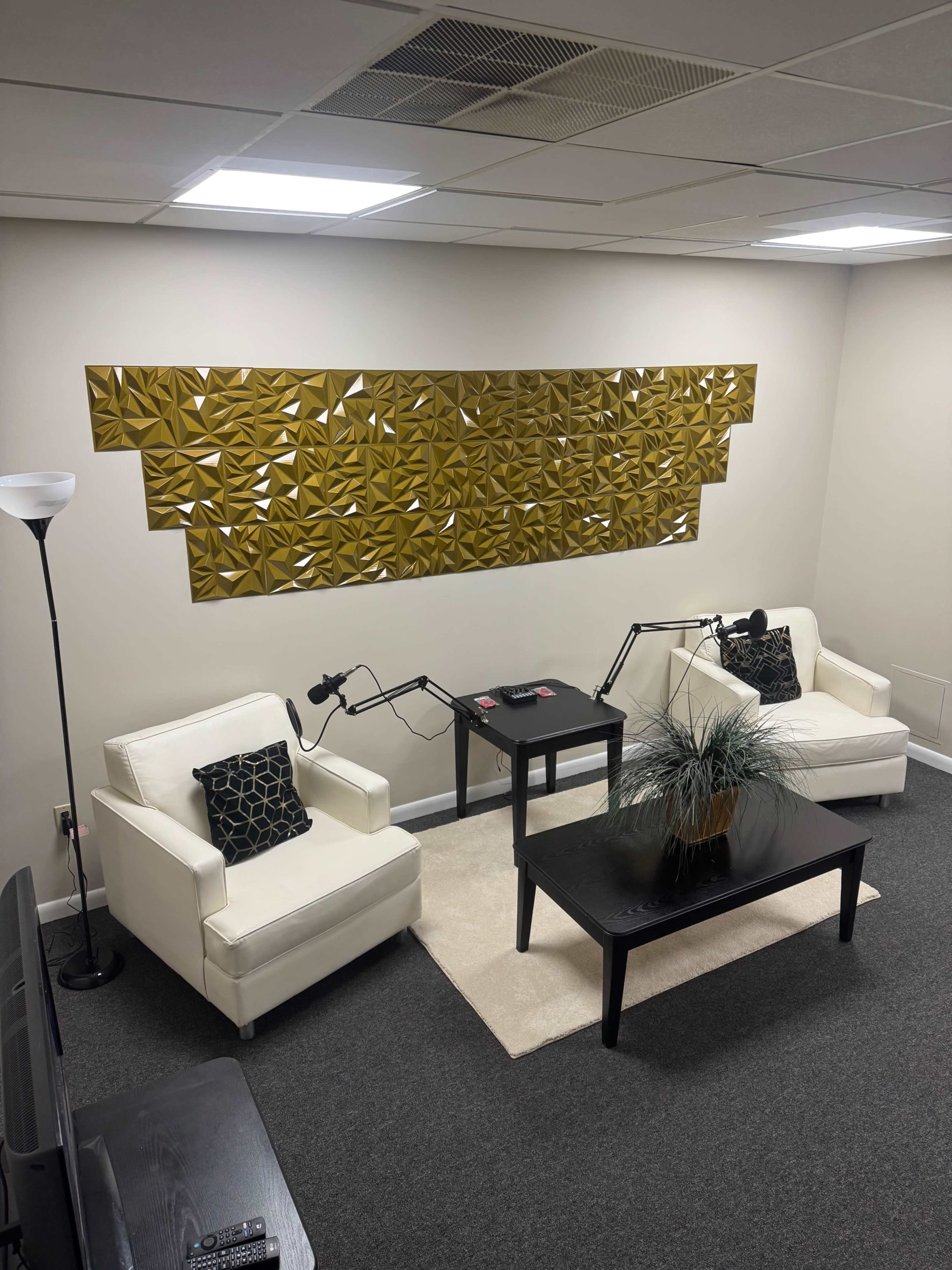 The image shows a small, modern lounge area featuring two white armchairs, a dark coffee table, and a decorative wall with geometric golden panels.