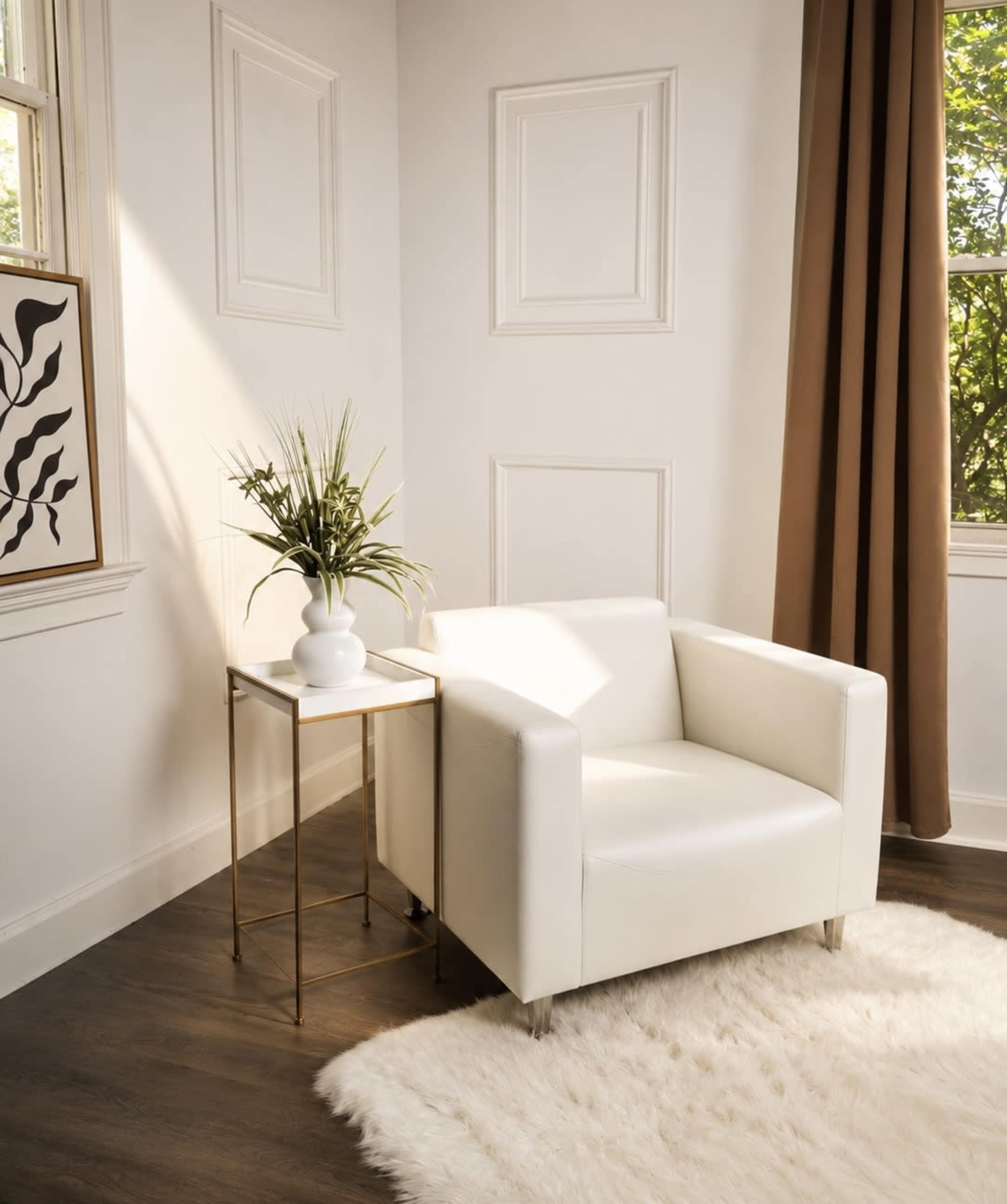 A white armchair is placed next to a small gold side table with a plant in a vase, situated in a room with natural light streaming through the windows.