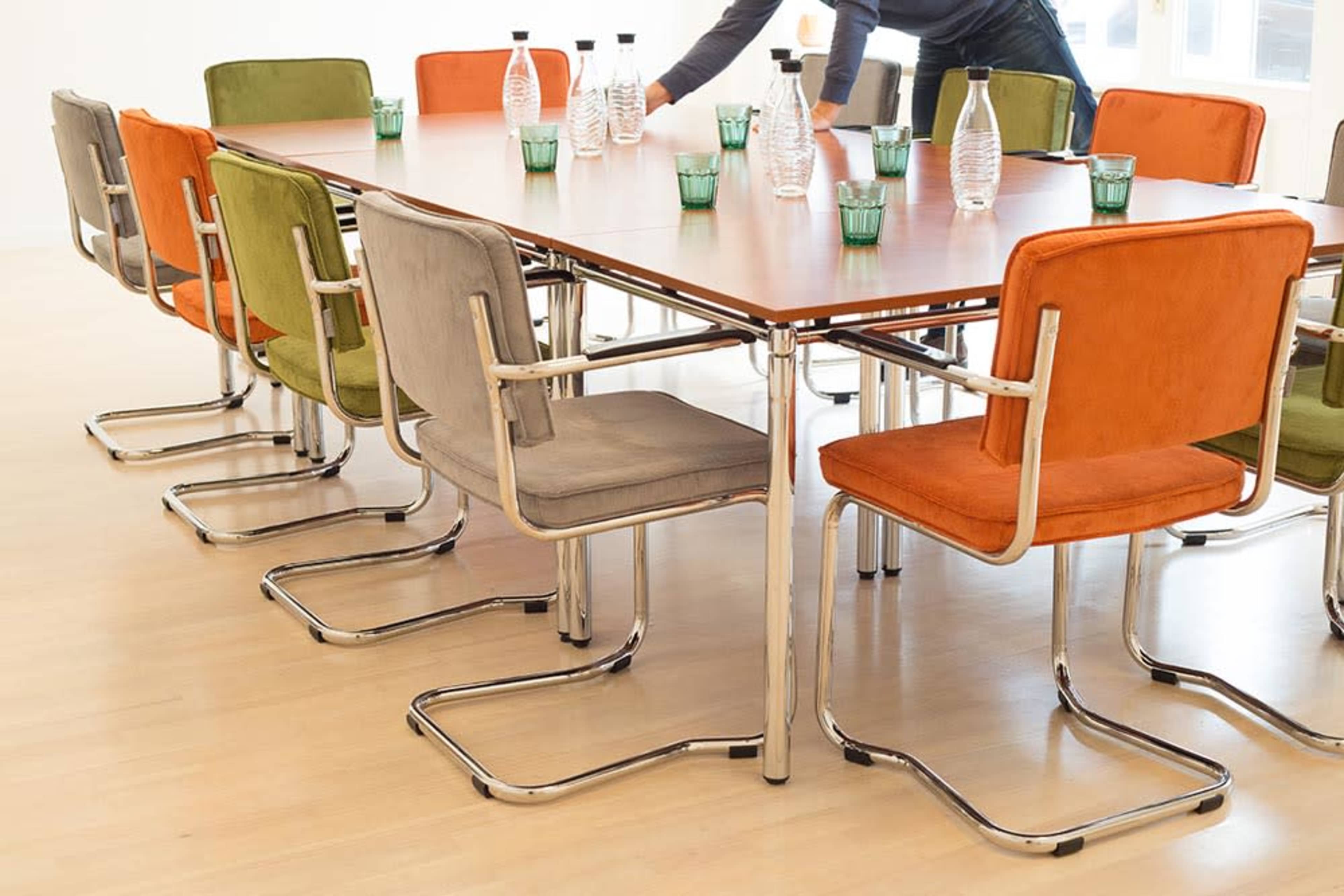 A rectangular conference table surrounded by colorful chairs, with water bottles and glasses arranged on top.