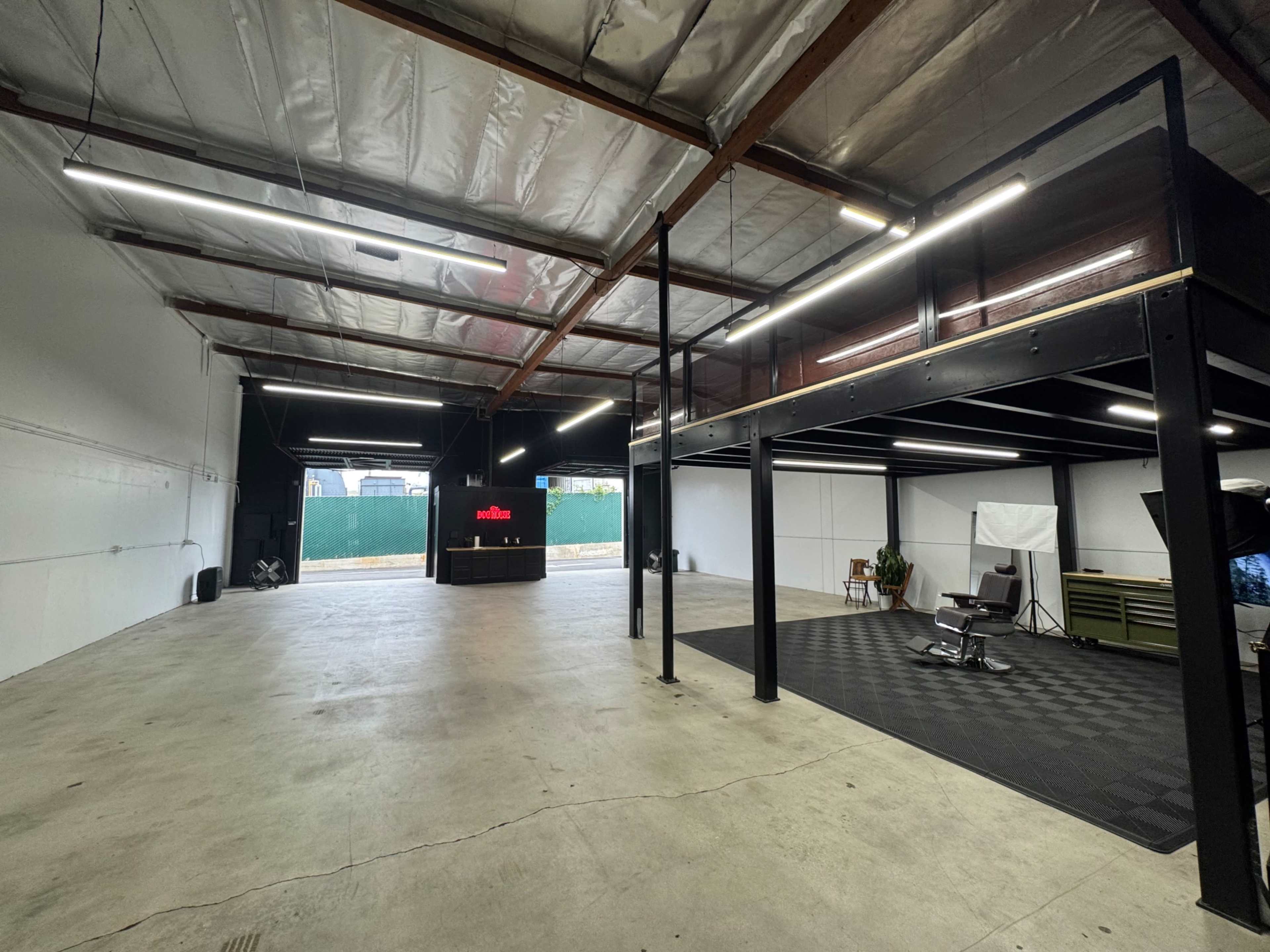 The image shows a spacious, empty warehouse with high ceilings, featuring a lower-level workspace, large open area, and a doorway leading to an outdoor space.