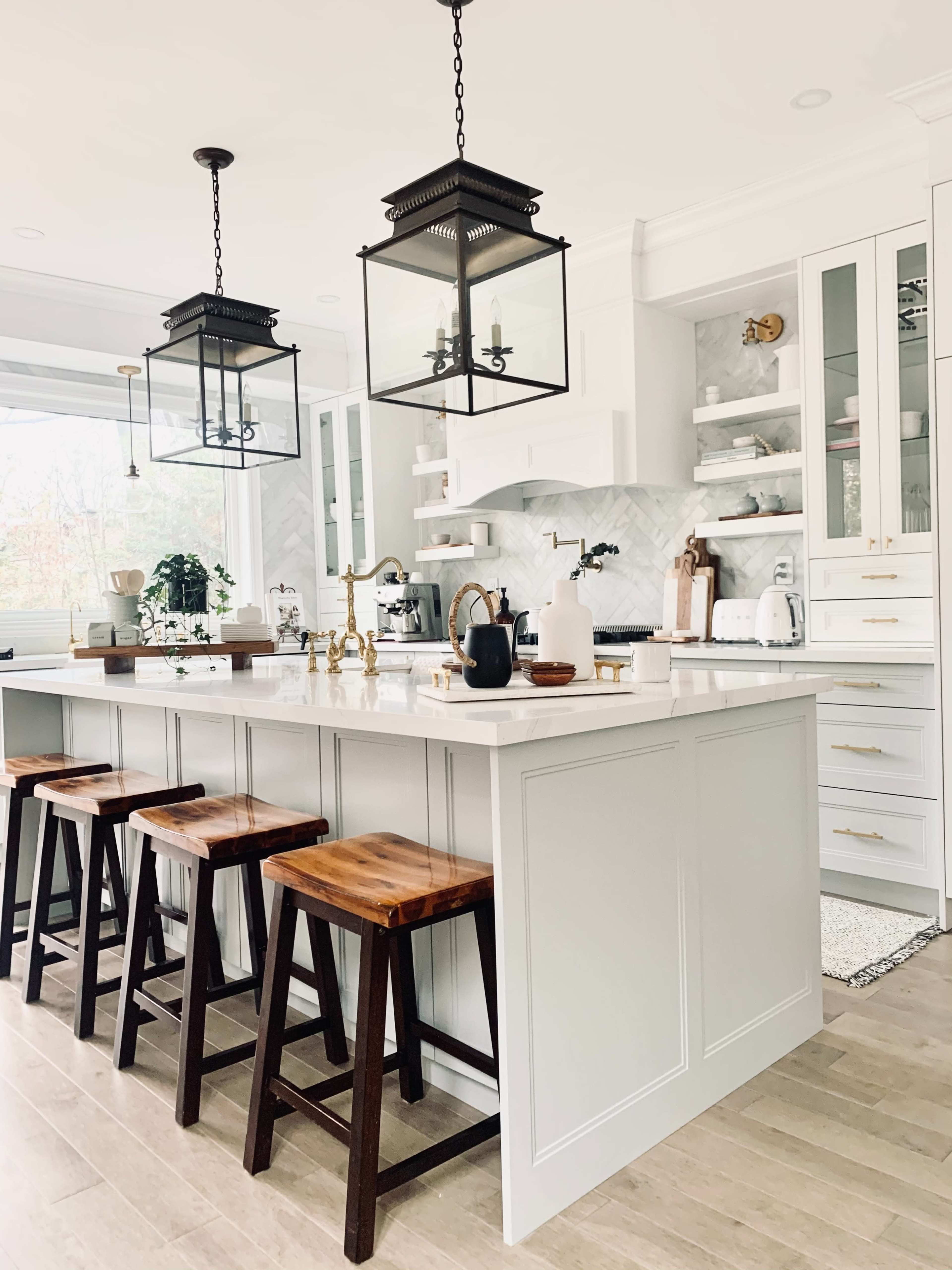 A modern kitchen features a central island with four wooden stools, pendant lights, and white cabinetry against a marble backsplash.
