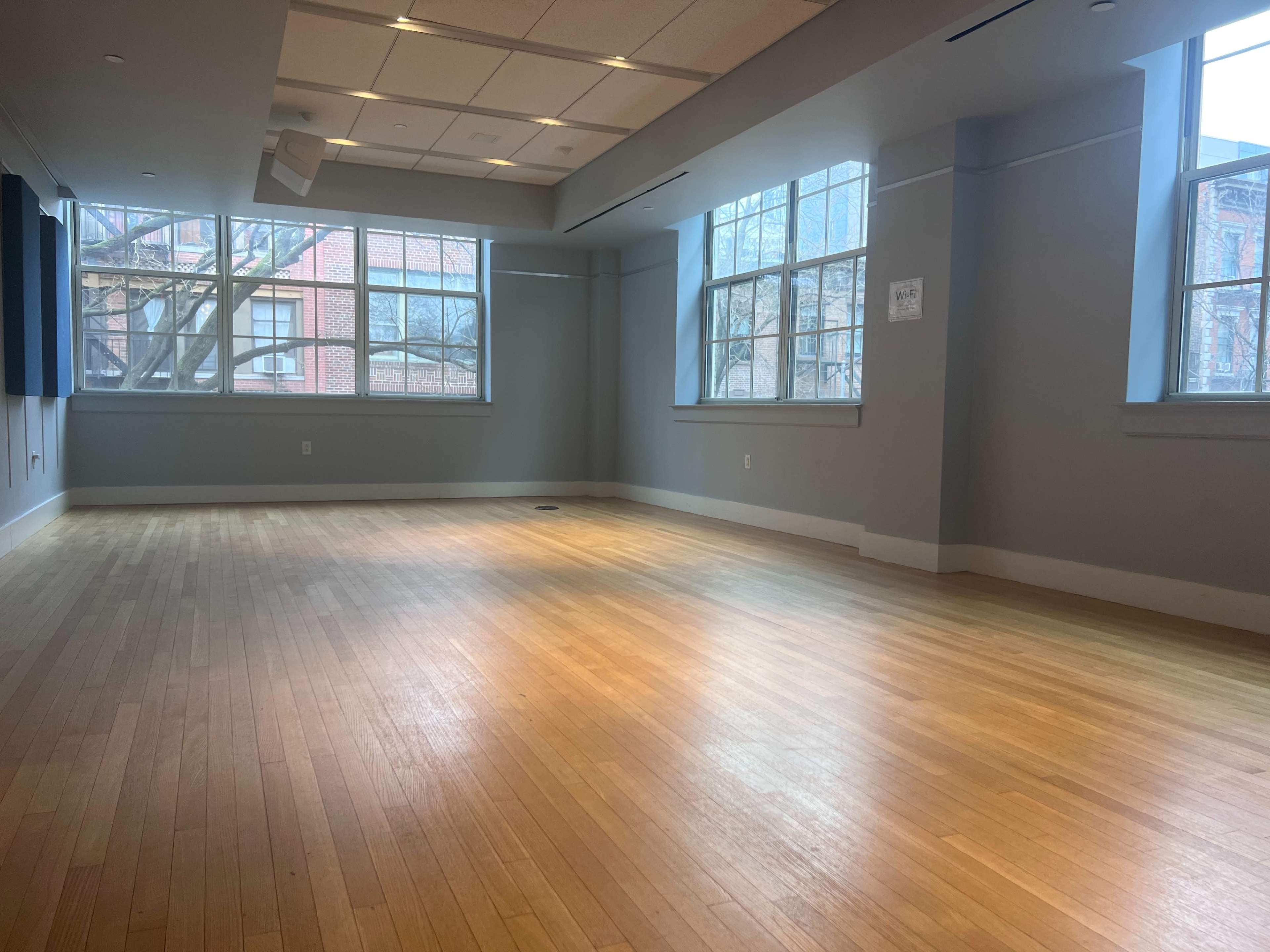 The image shows a spacious, empty room with hardwood floors and large windows letting in natural light.