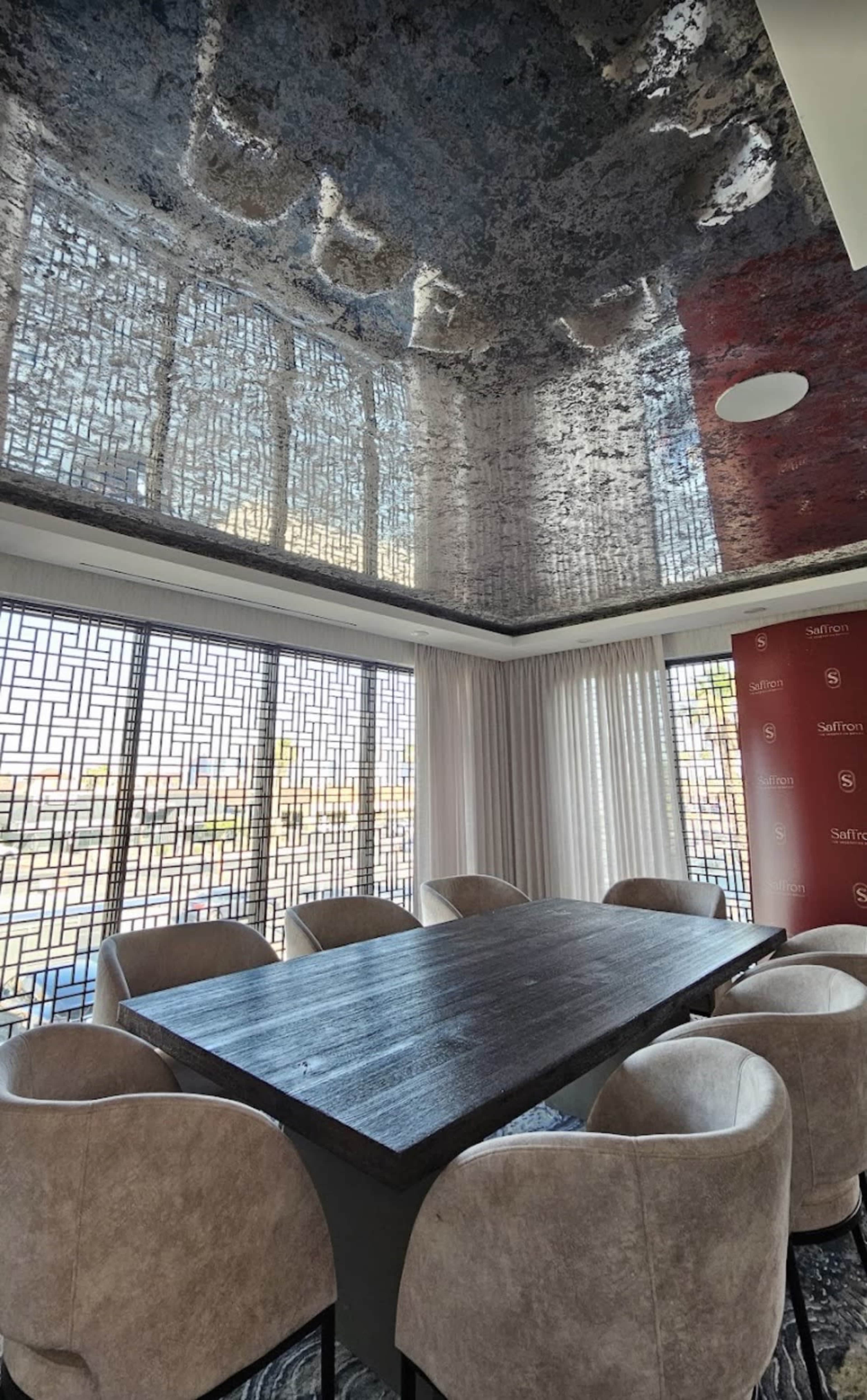 A large conference room features a dark wooden table surrounded by beige upholstered chairs, with a reflective ceiling and geometric window designs.