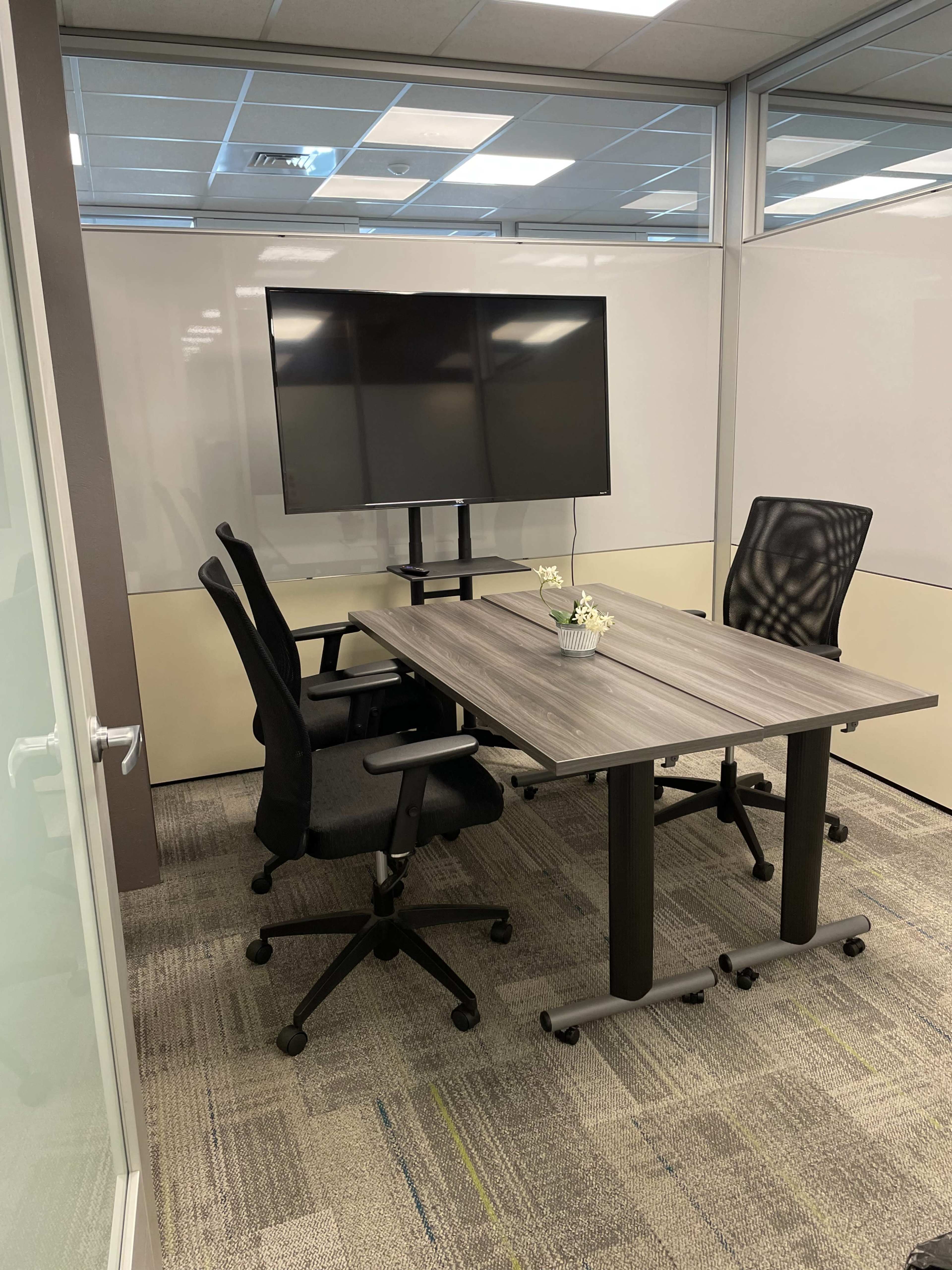 The image shows a small meeting room with a wooden table, two office chairs, and a wall-mounted television.