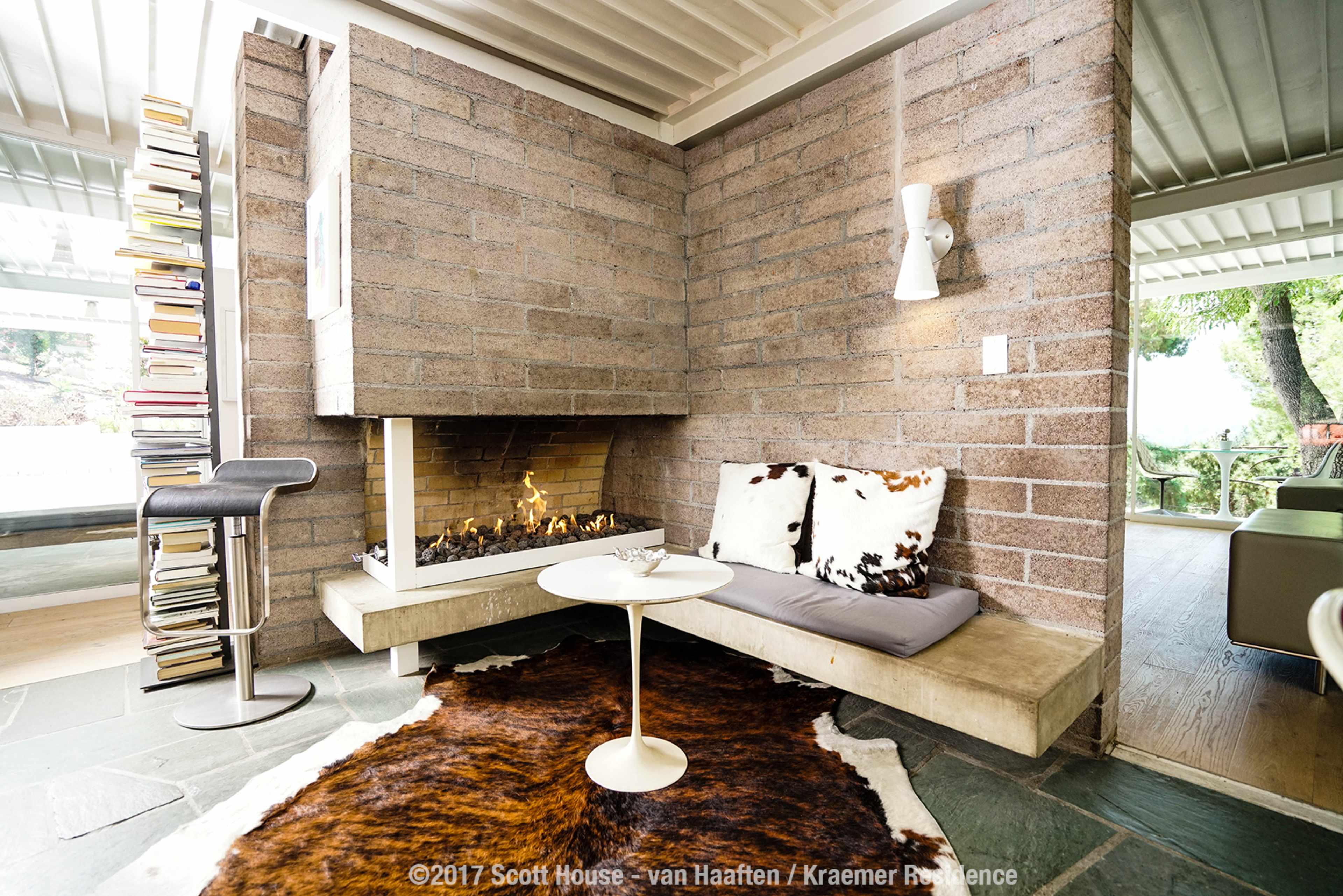 A modern living space featuring a stone fireplace, a corner seating area with a small table, a bookshelf, and natural light filtering in from large windows.