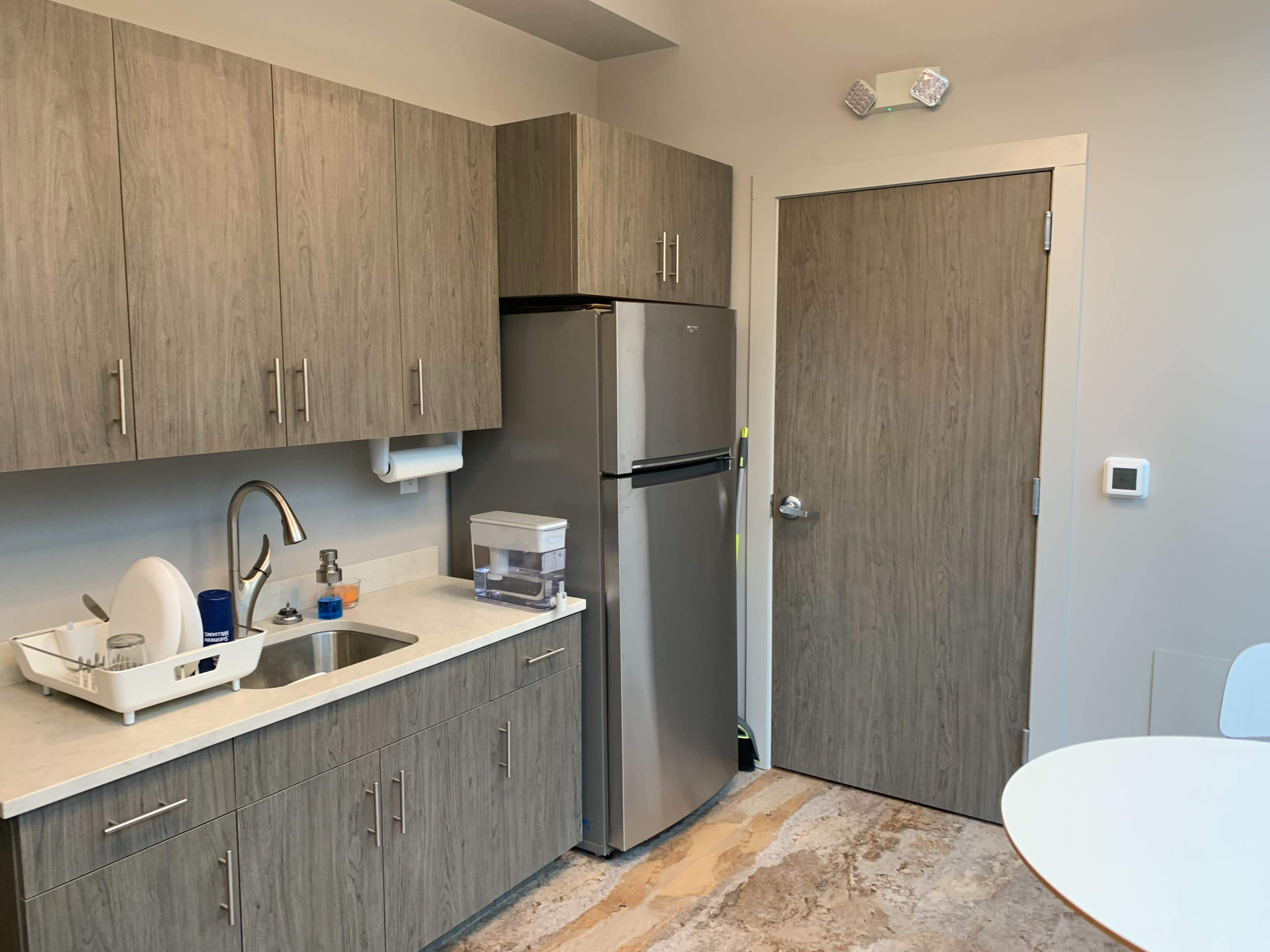A modern kitchen area featuring gray cabinets, a stainless steel refrigerator, a sink, and a door leading to another room.