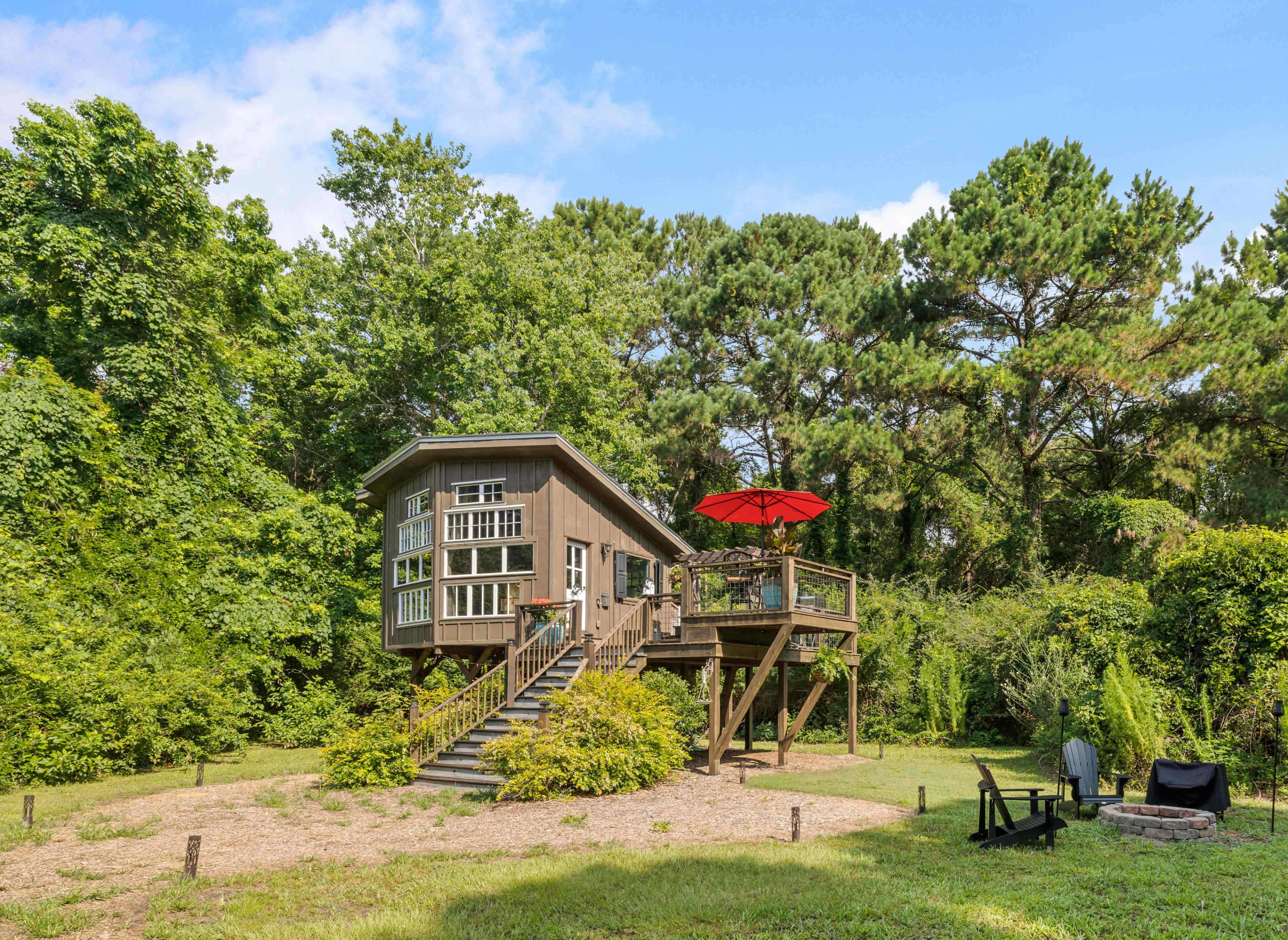 A two-story house with large windows and a red umbrella on the deck is situated on a grassy area surrounded by trees.