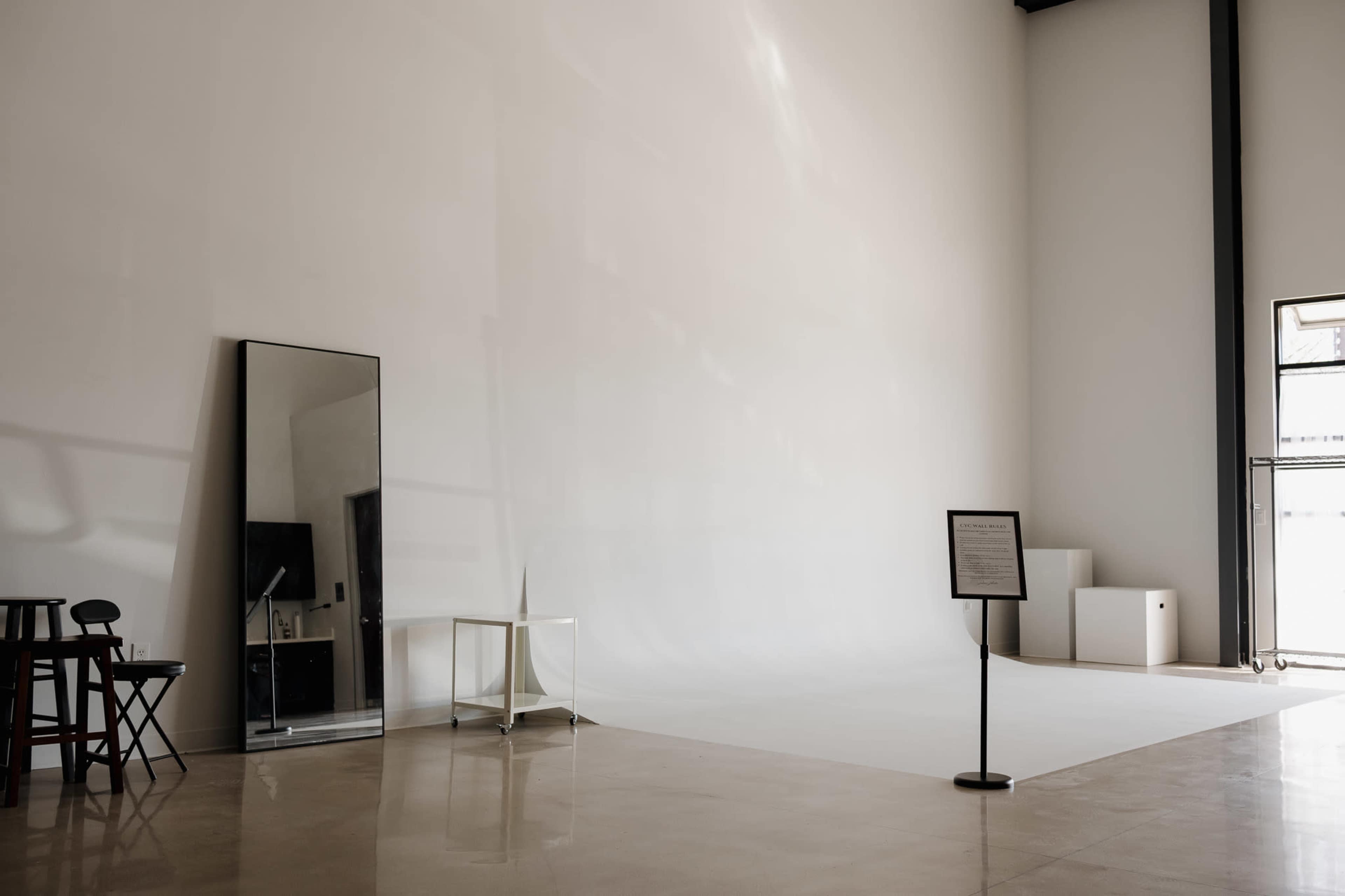 A spacious, minimalist studio features a large white wall, a full-length mirror, a table, and some small boxes in the corner.