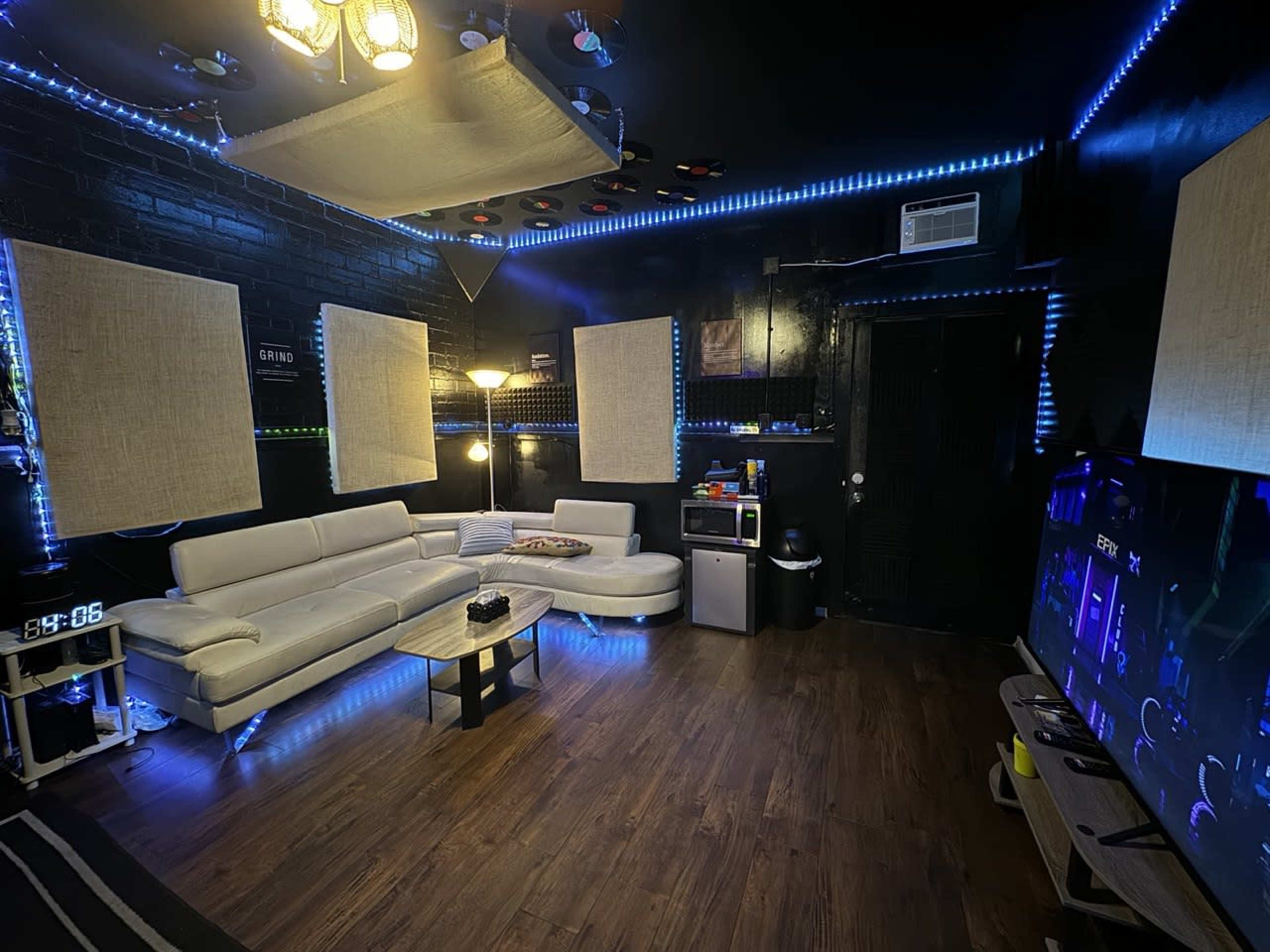 A modern, dimly lit lounge features a white sectional sofa, a coffee table, and a large TV against a wall illuminated by blue LED lights.