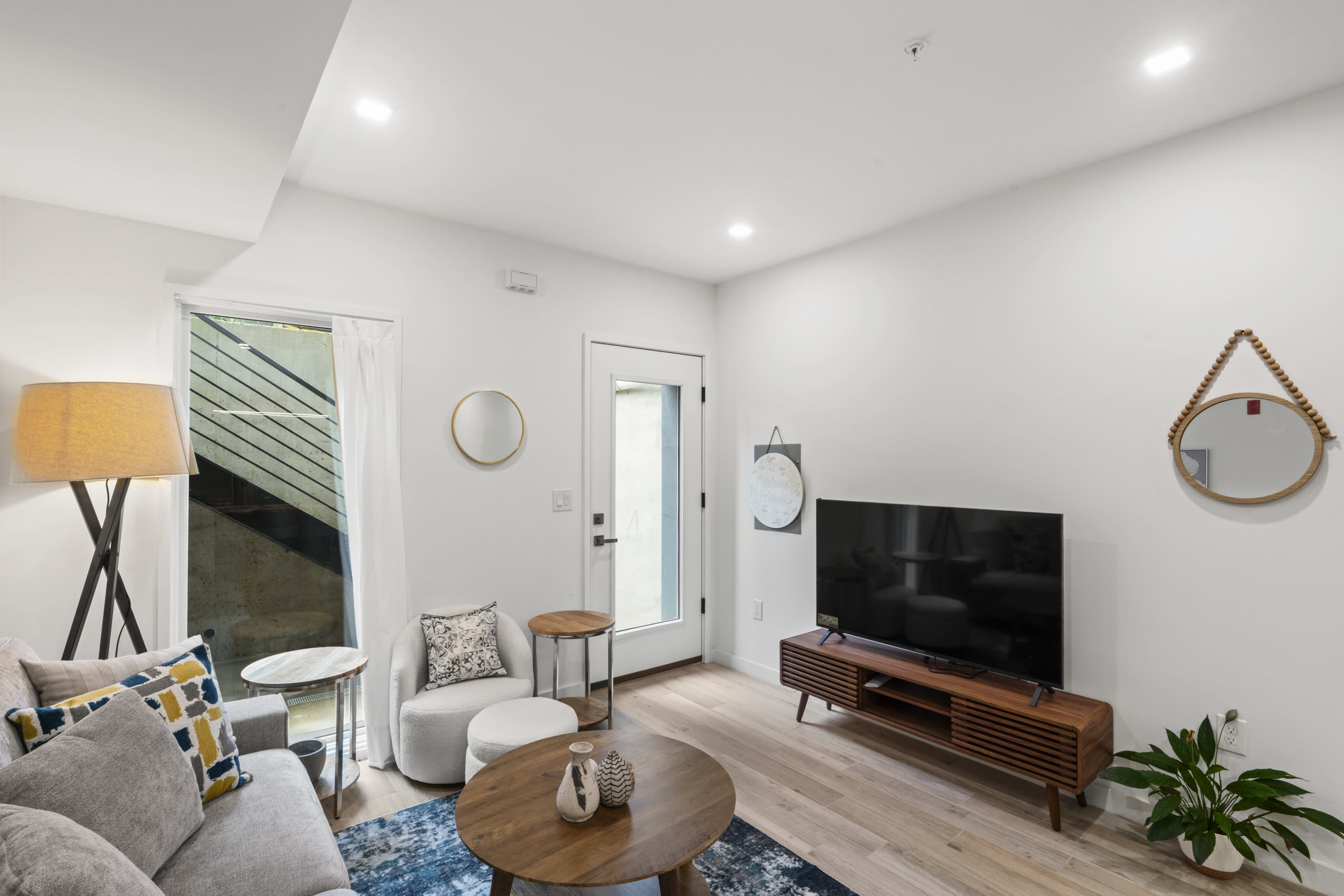 The image shows a modern living room featuring a gray sofa, a coffee table, a television stand with a TV, a round mirror, and a doorway leading outside.