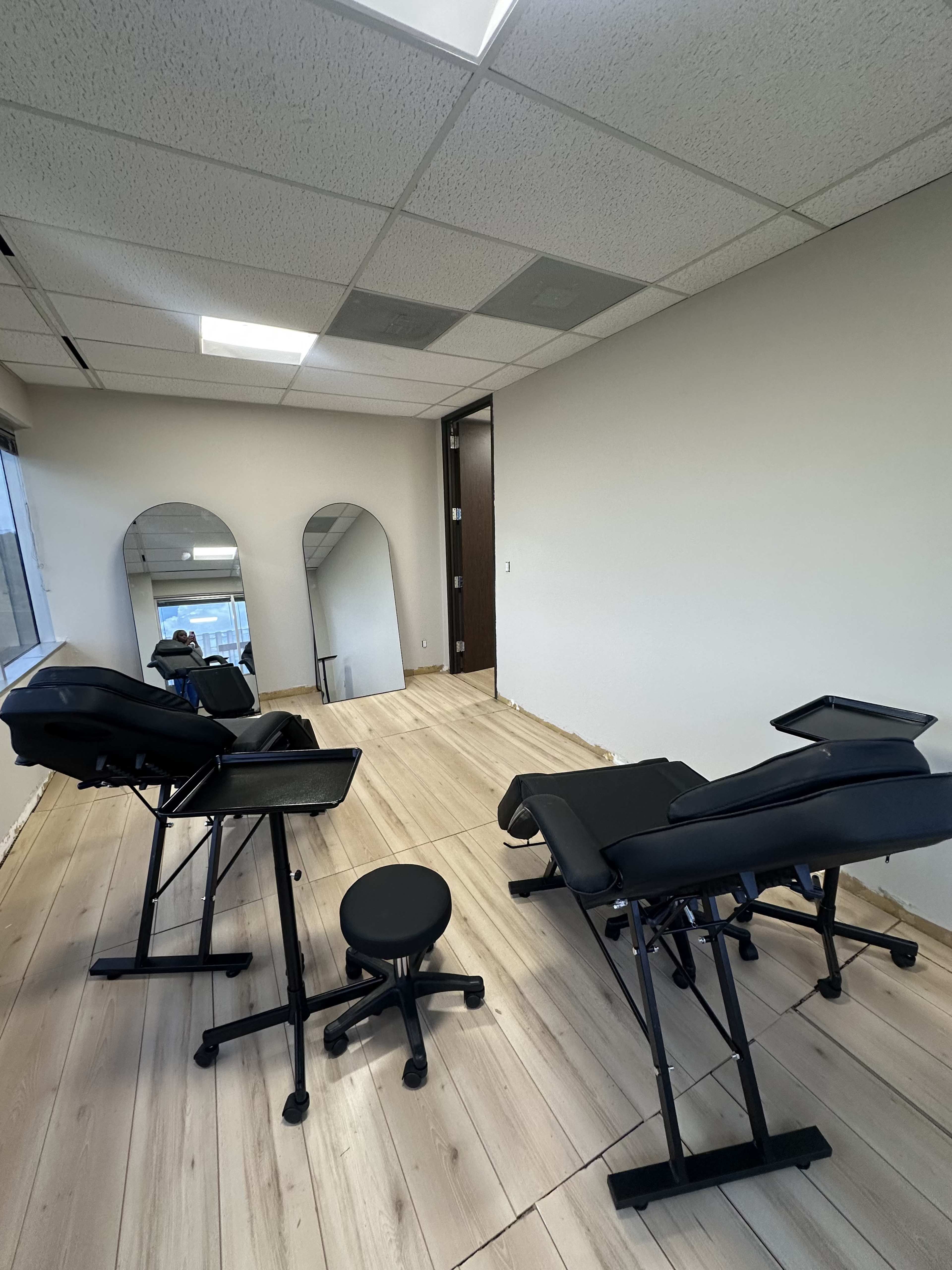 The image shows a partially furnished room with two black massage tables, a black stool, and wood flooring, along with a large window and an open doorway.