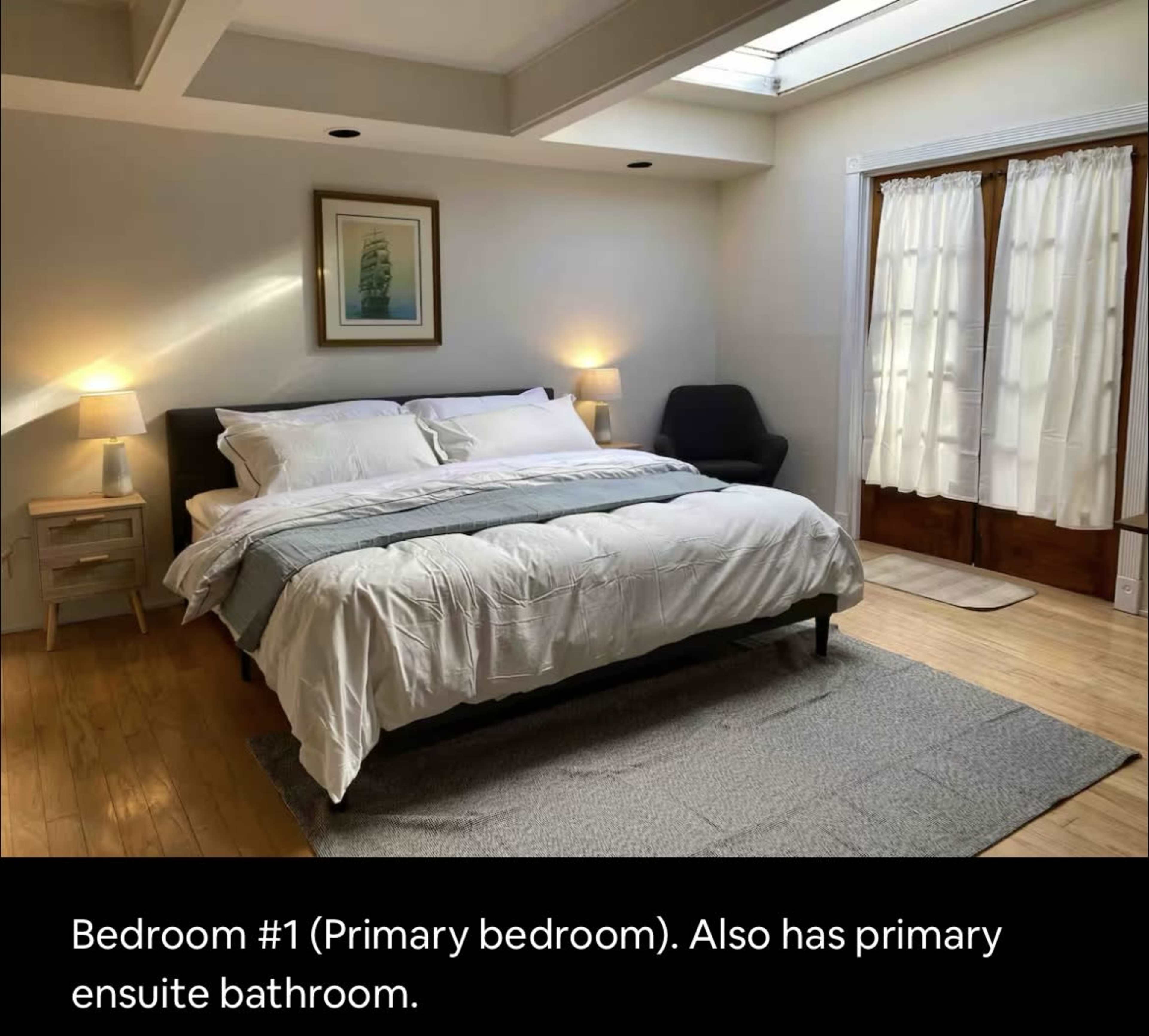 A primary bedroom features a large bed with white bedding, bedside lamps, and a skylight, along with doors leading to a bathroom.