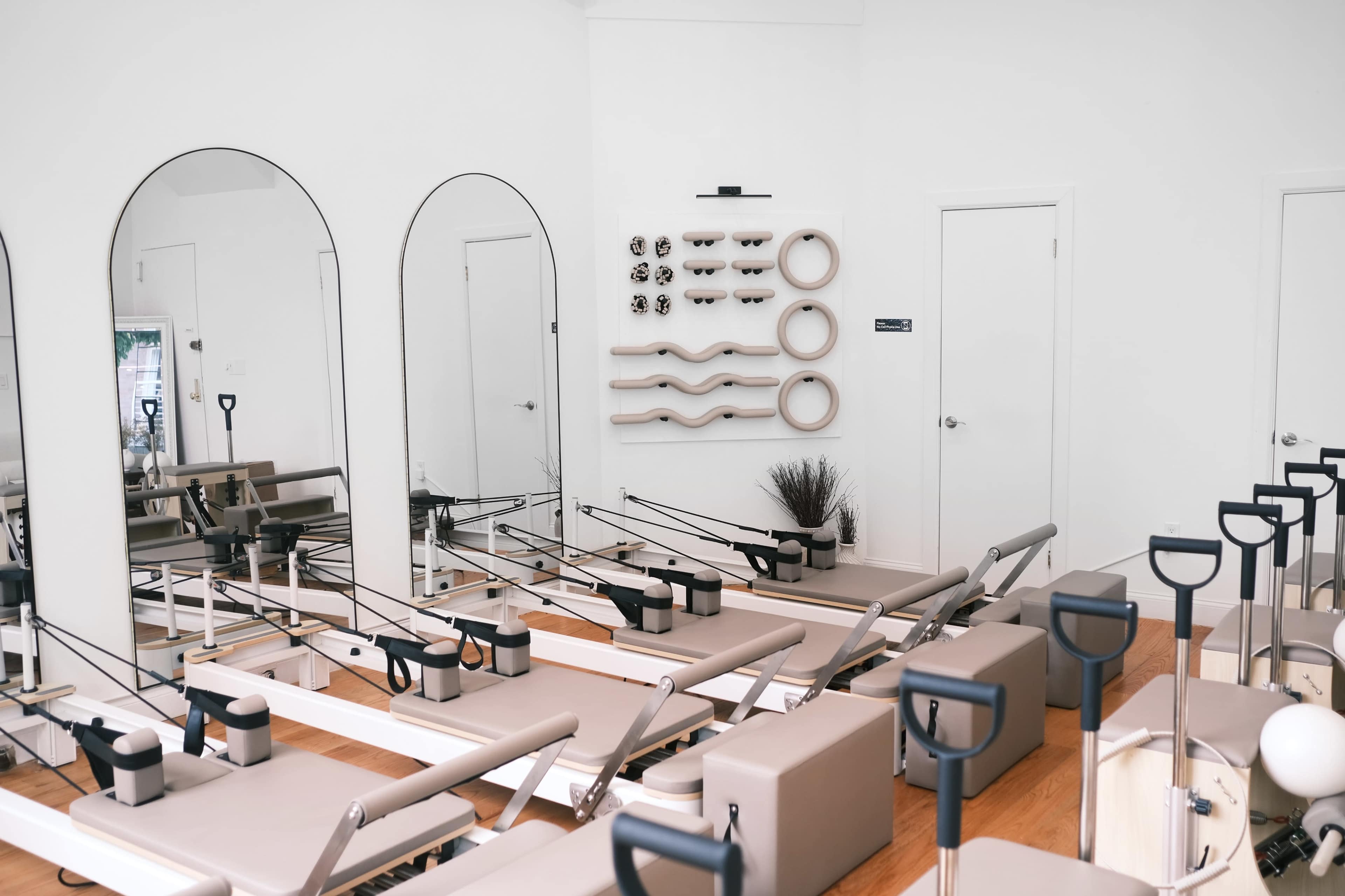 The image shows a minimalist Pilates studio featuring several reformers, mirrors on the walls, and a decorative panel with wavy shapes.