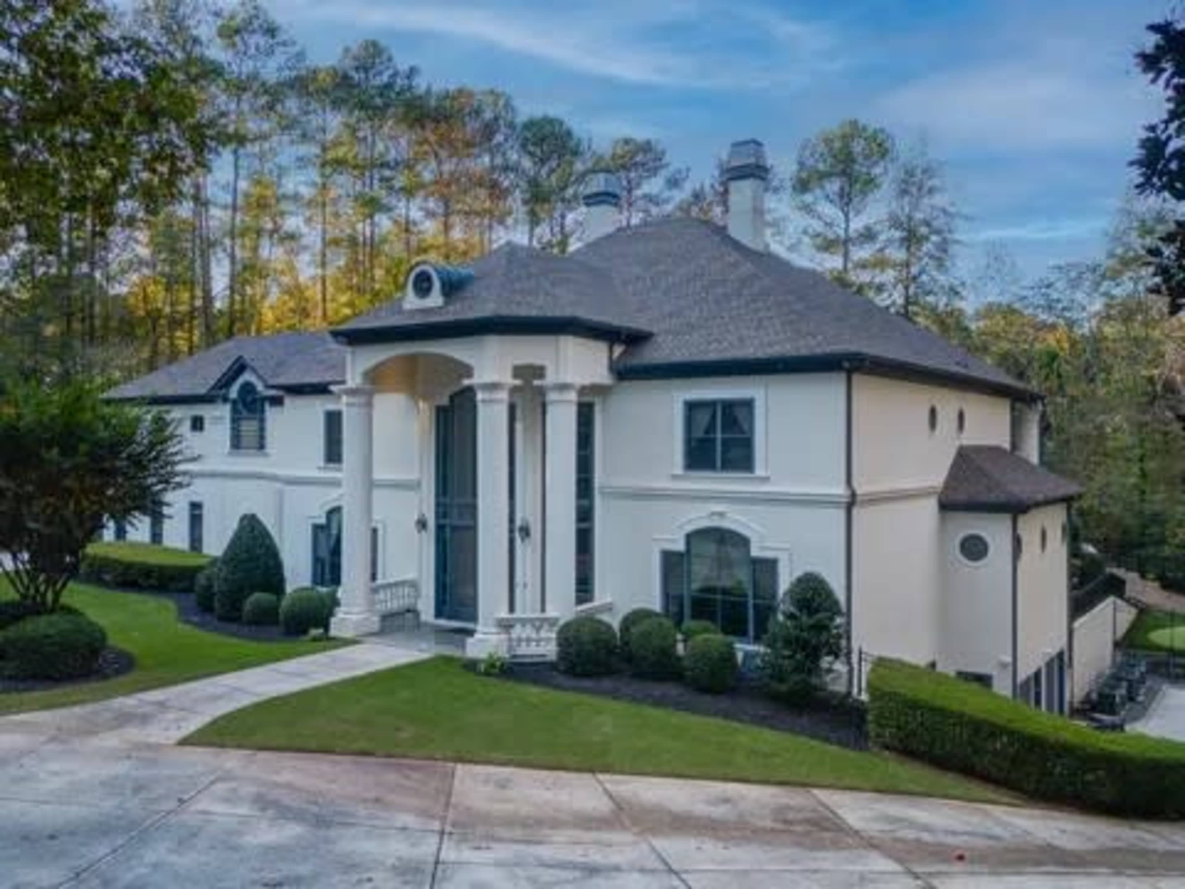 The image shows a large, modern home with a white exterior, prominent columns at the entrance, and landscaped greenery surrounding the property.