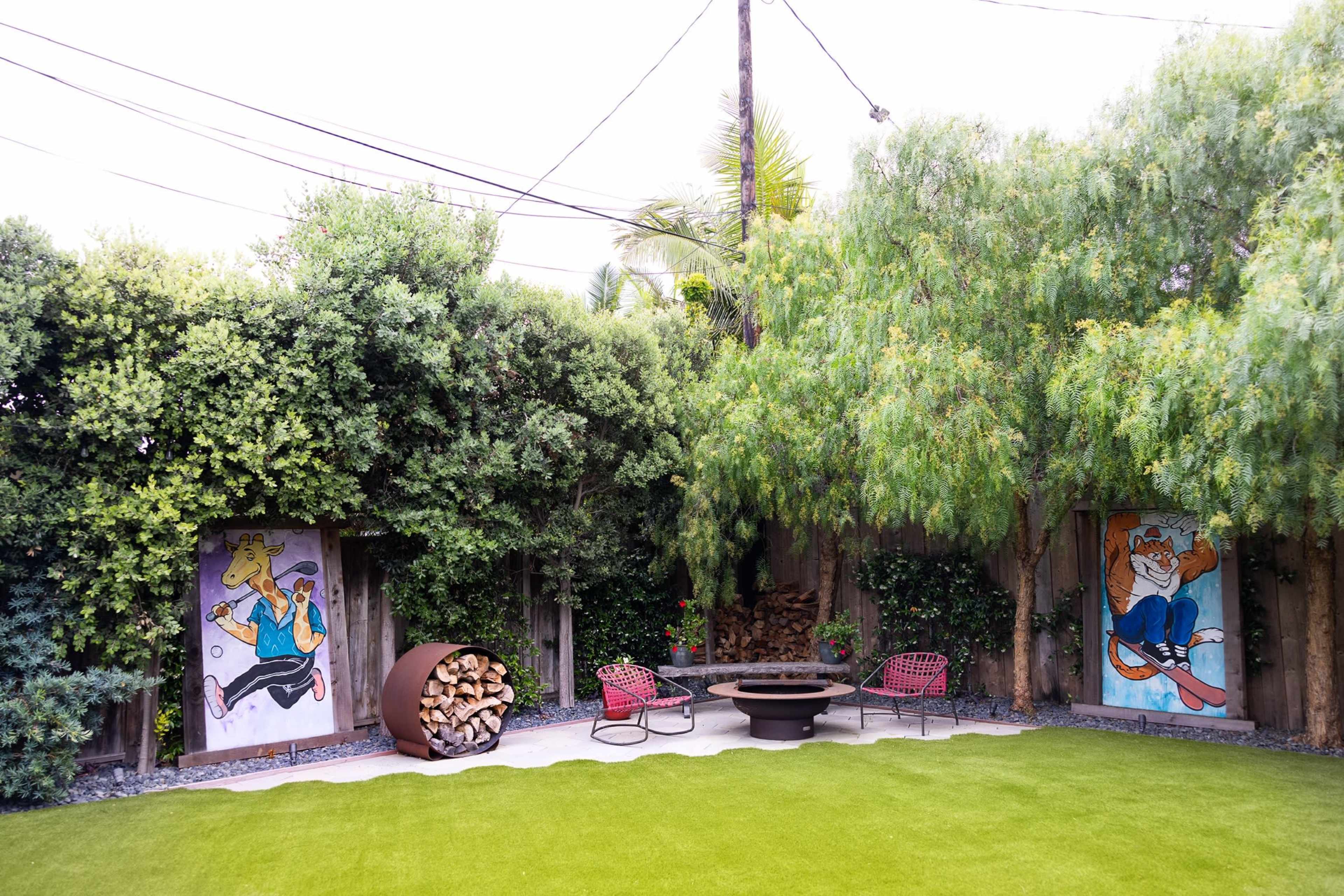 The image shows a backyard featuring a grassy area, a circular fire pit, two red chairs, and two large murals on wooden fences.