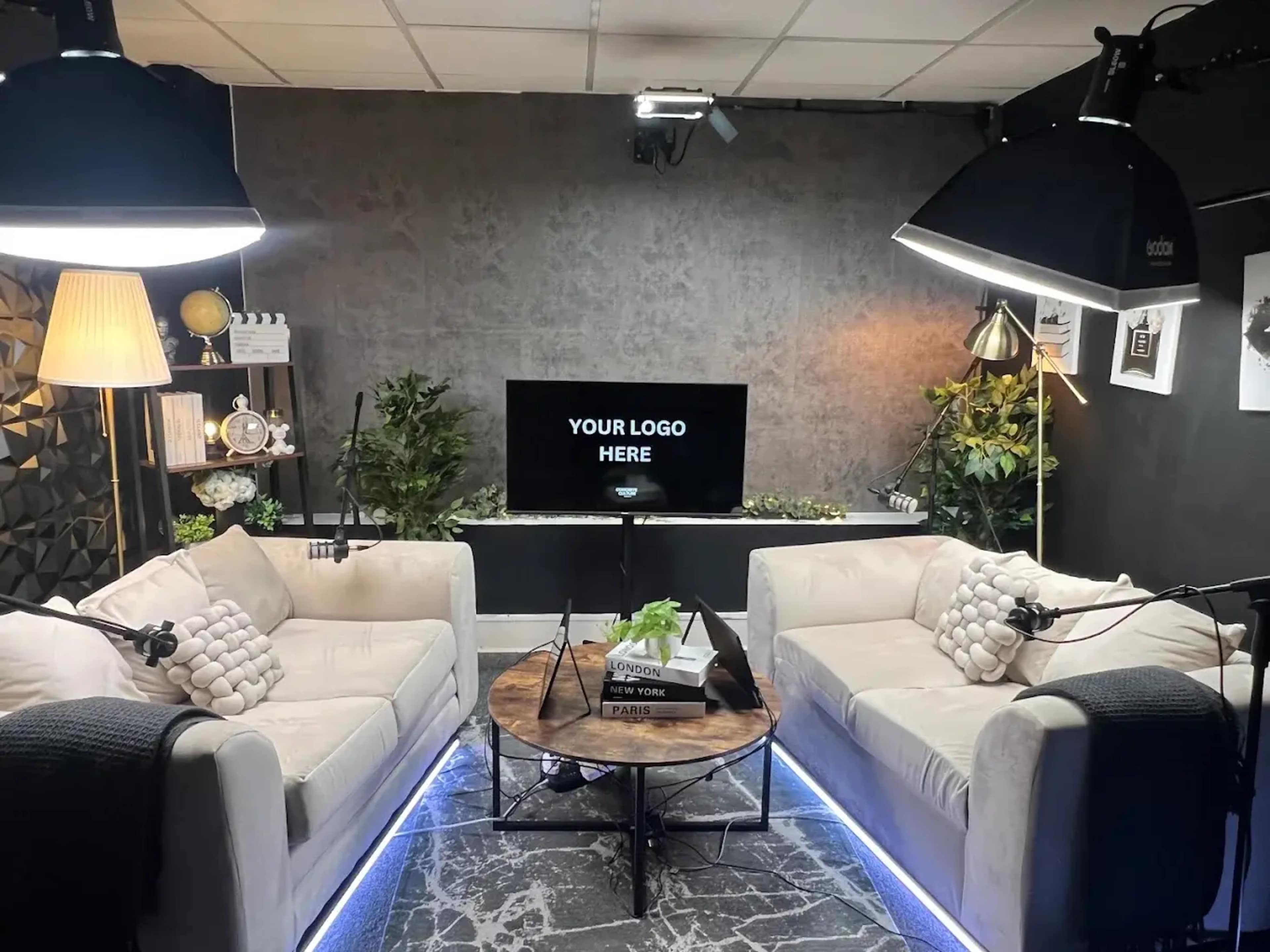 A well-lit modern studio space features two sofas, a round coffee table, and a screen displaying "YOUR LOGO HERE" against a textured wall.