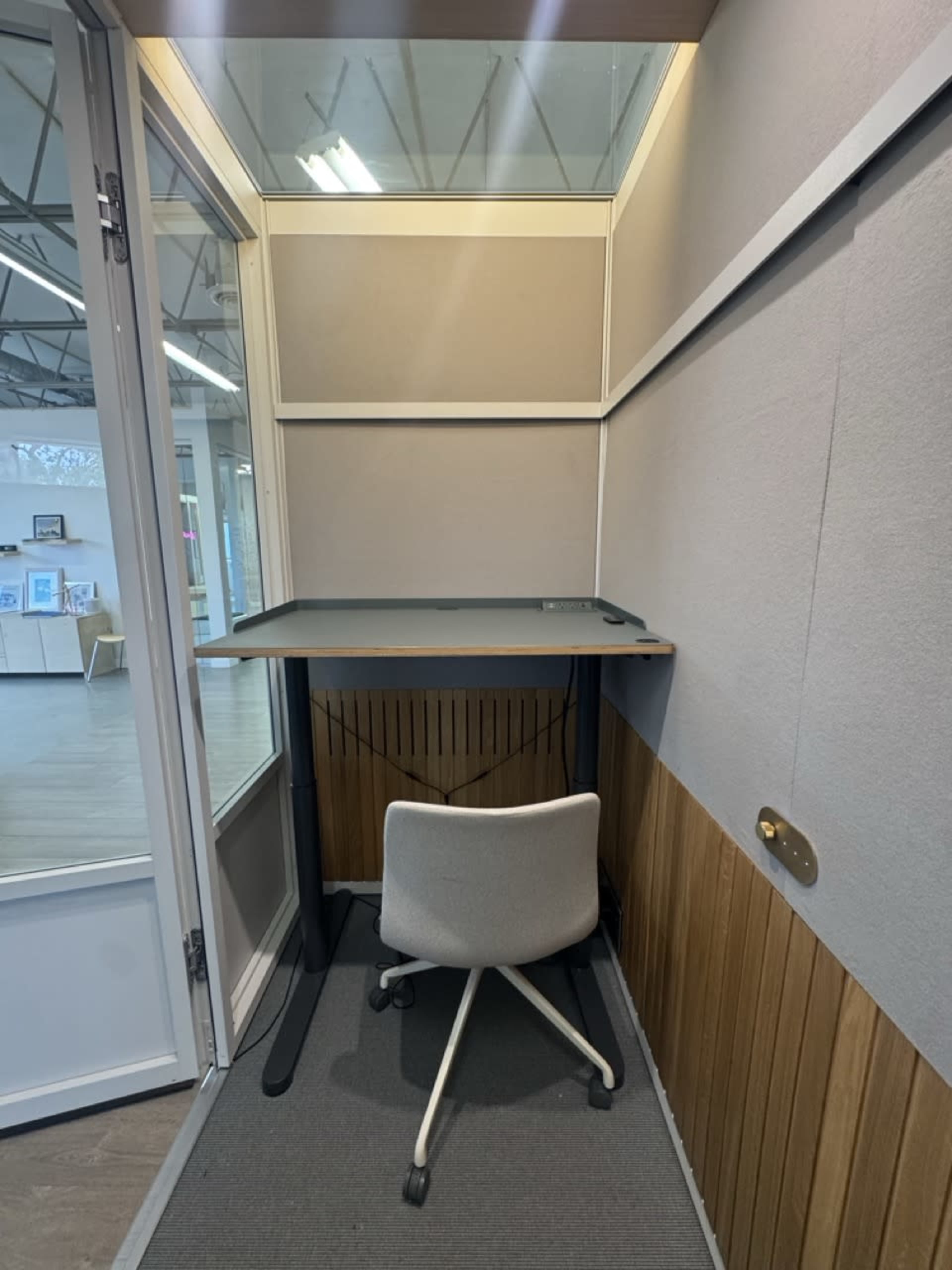 The image shows a small, enclosed workspace featuring a simple desk and a wheeled chair against a wall, with a glass door visible on one side.
