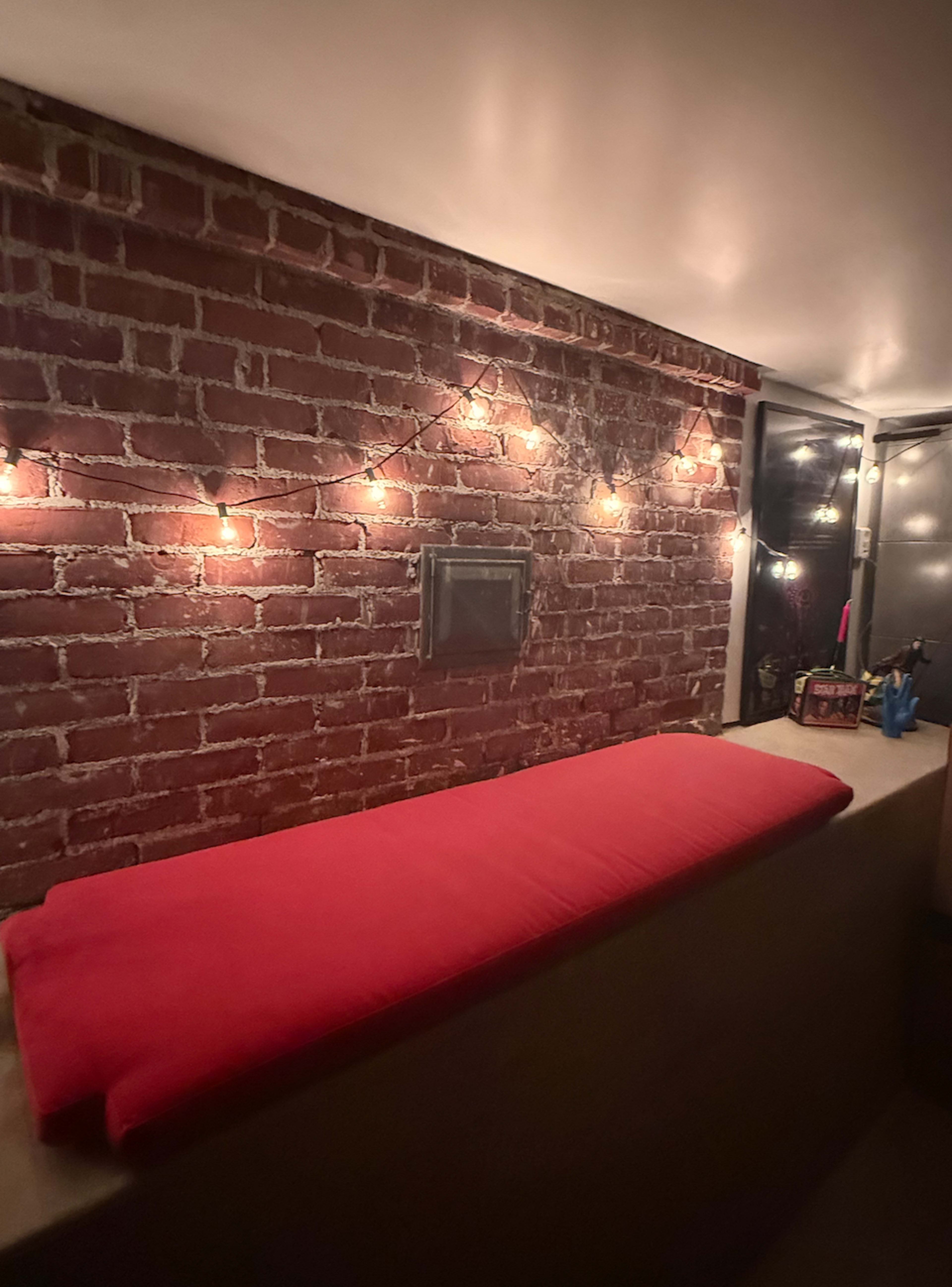 The image shows a cozy nook with a red cushion on a wooden ledge against a brick wall, accented by string lights.