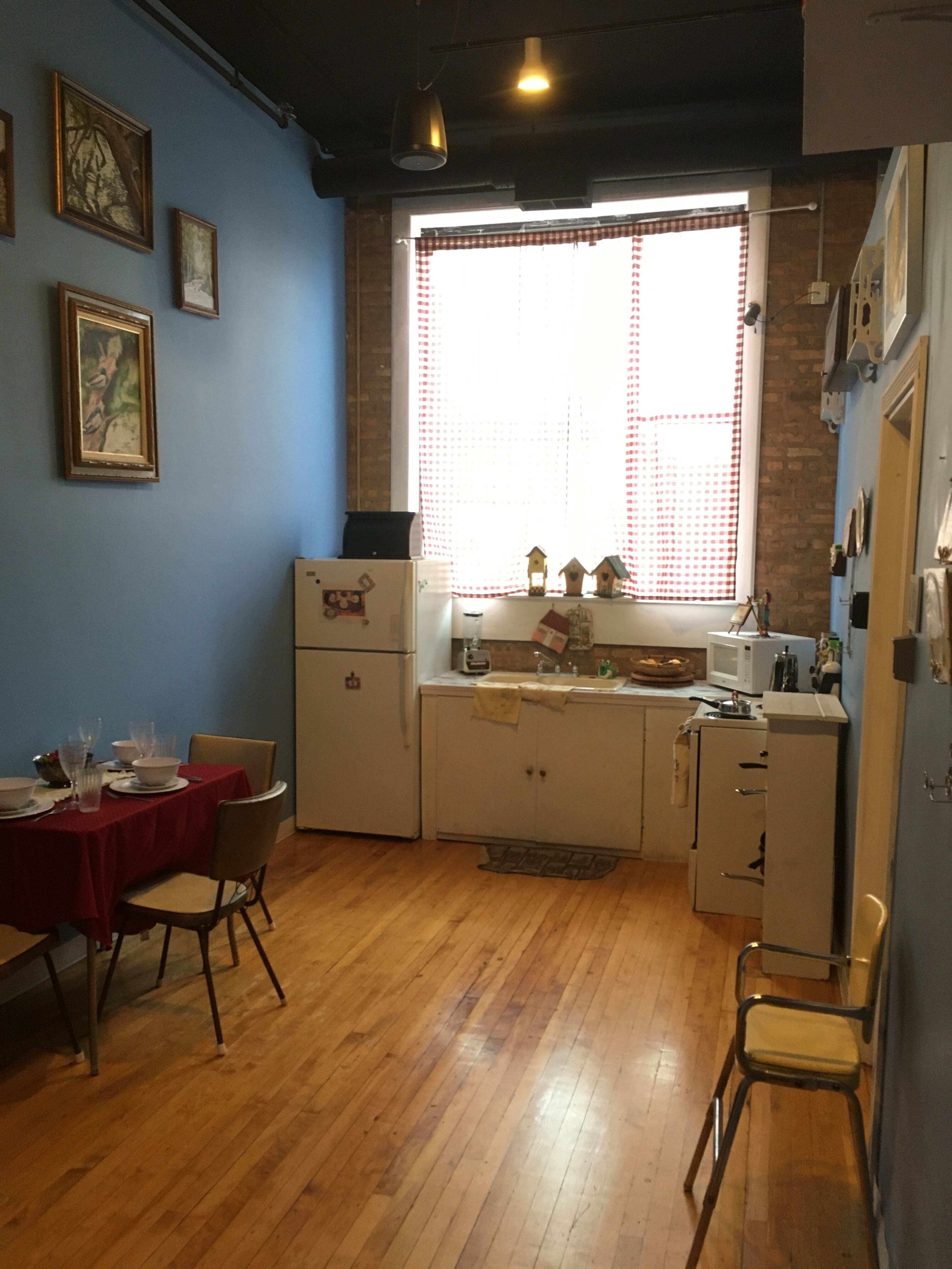 A small kitchen space with a red table, chairs, a refrigerator, and various kitchen appliances against a blue wall, featuring framed artwork and a large window.