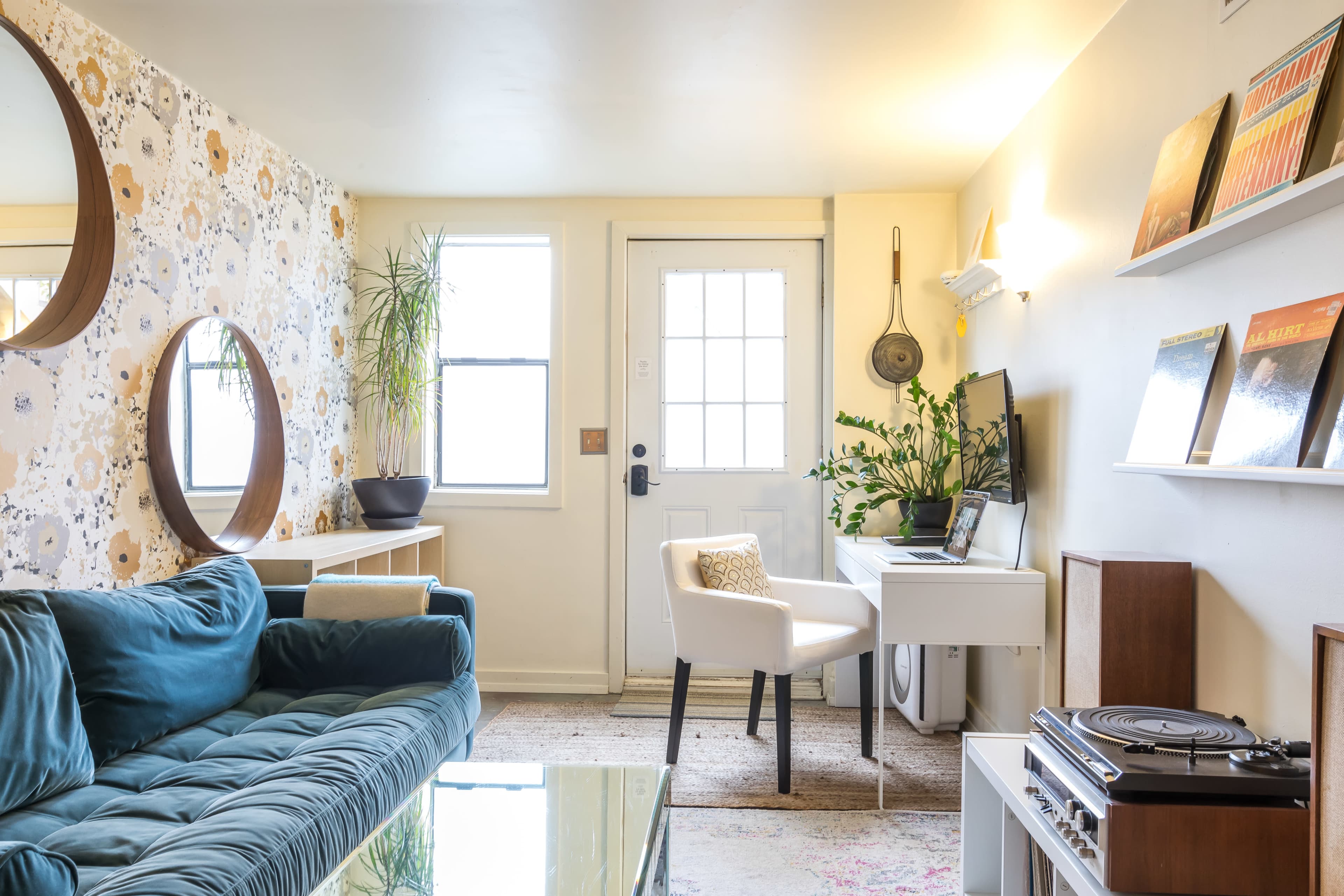 A cozy living room features a teal sofa, a small white desk with a computer, and a door leading outside, surrounded by decorative plants and patterned wallpaper.