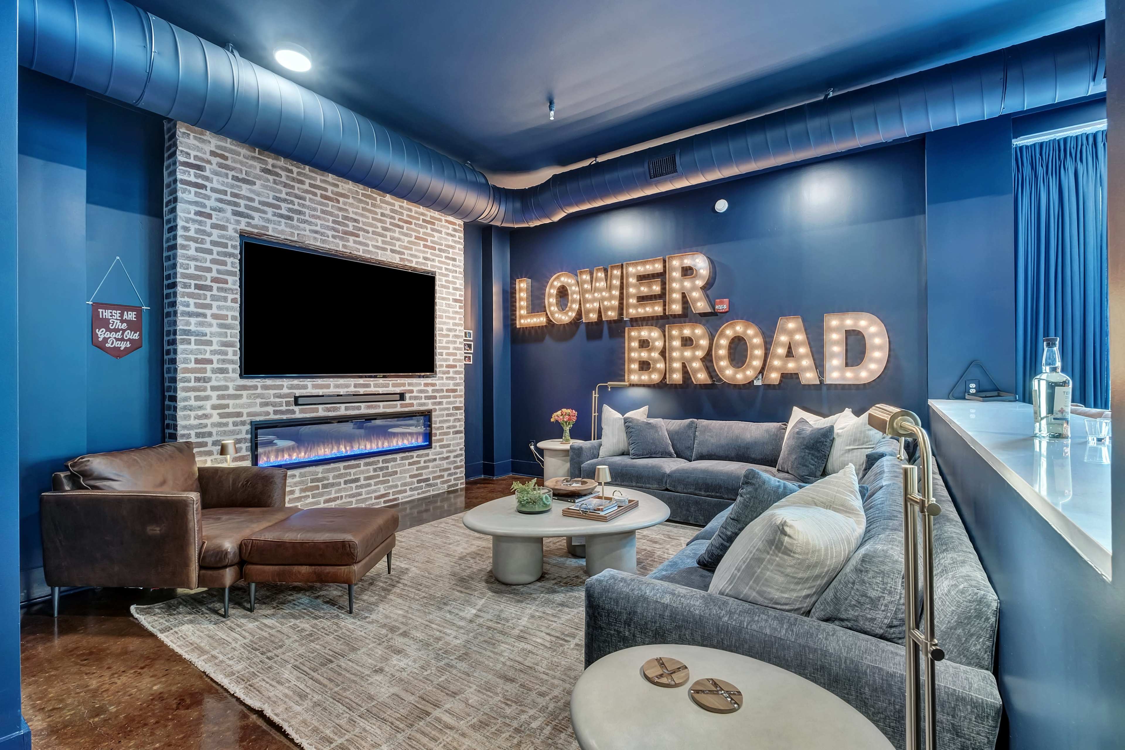 The image shows a modern living room with a sectional sofa, two armchairs, a coffee table, and a large wall featuring illuminated letters spelling "LOWER BROAD" alongside a television and a decorative fireplace.