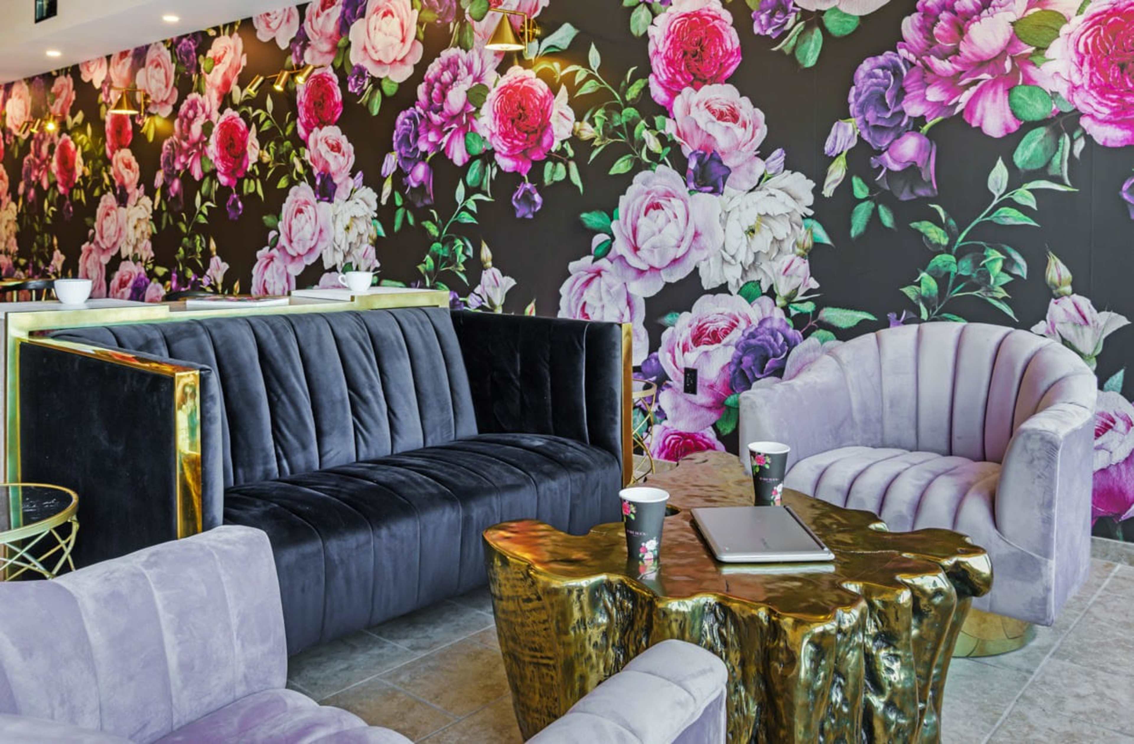 The scene features a modern seating area with a black velvet sofa, two light purple armchairs, and a gold-accented coffee table, set against a floral wallpaper of large pink and white roses.
