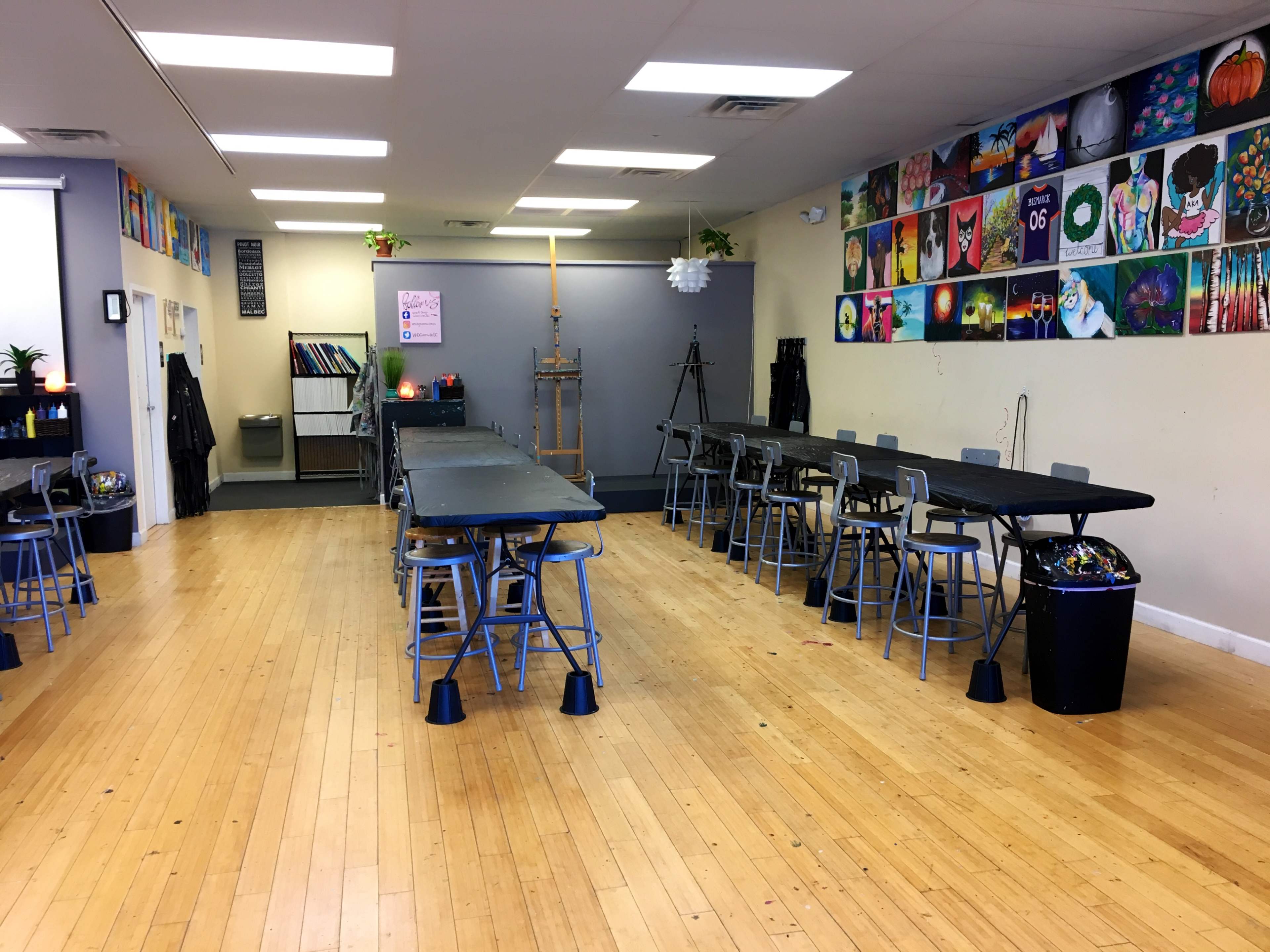 A spacious art studio with multiple tables and chairs arranged neatly, and walls adorned with colorful paintings.