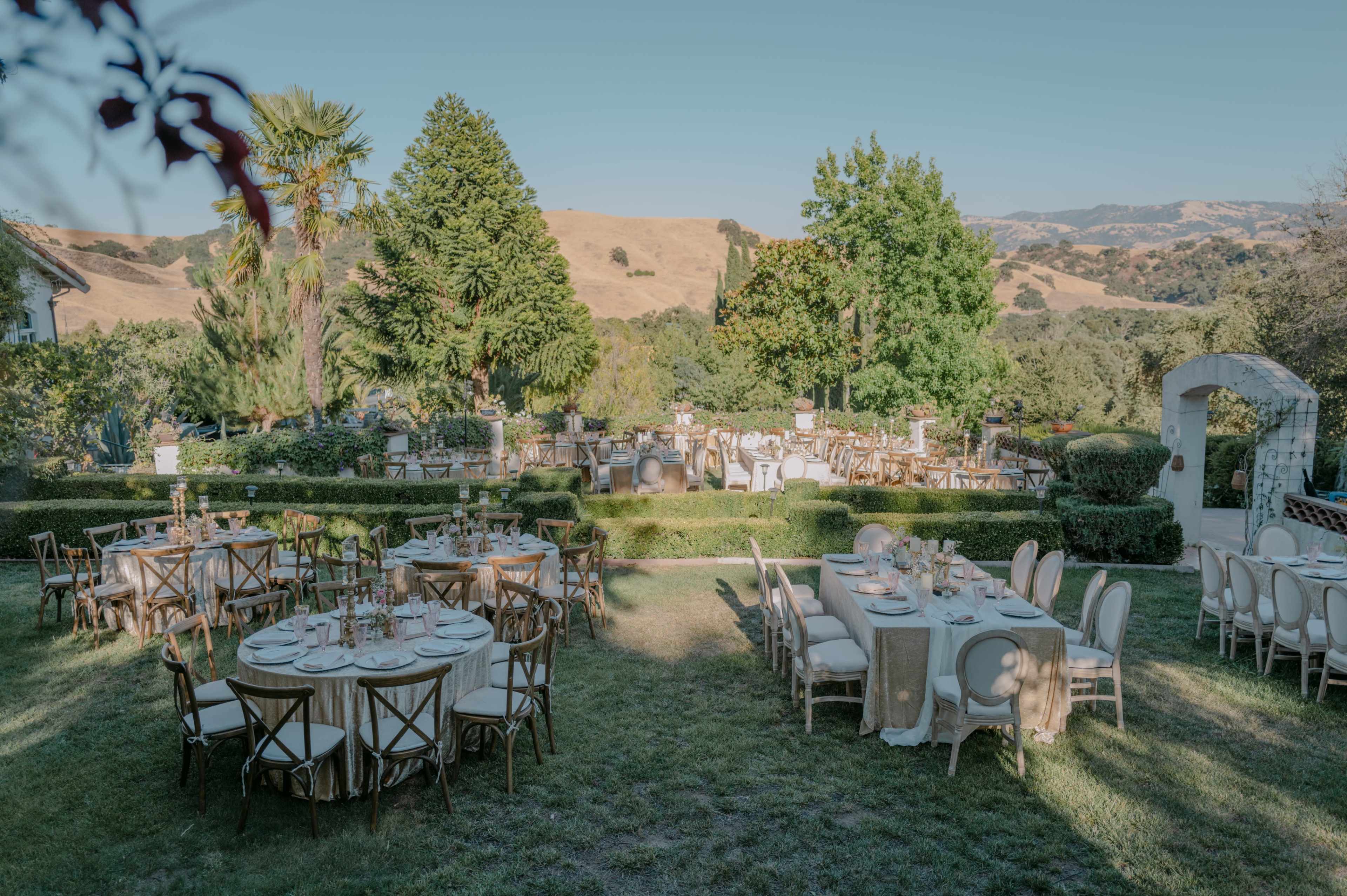 The image shows a beautifully arranged outdoor venue with several round and rectangular dining tables set up on green grass, surrounded by trees and hills in the background.