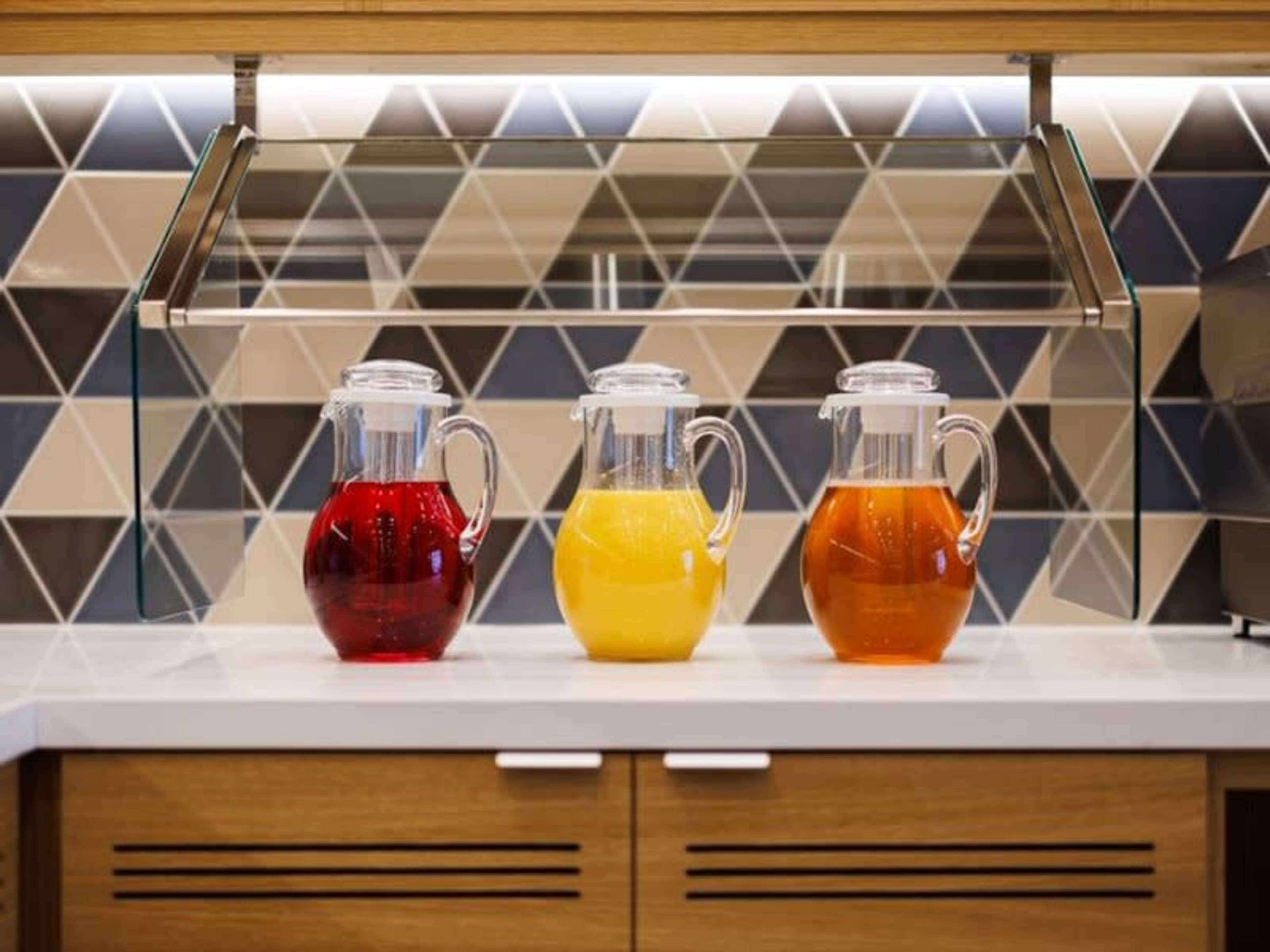 Three glass pitchers filled with red, yellow, and orange beverages are lined up on a white countertop beneath a glass cover, with a geometric tiled backsplash in shades of blue and beige.