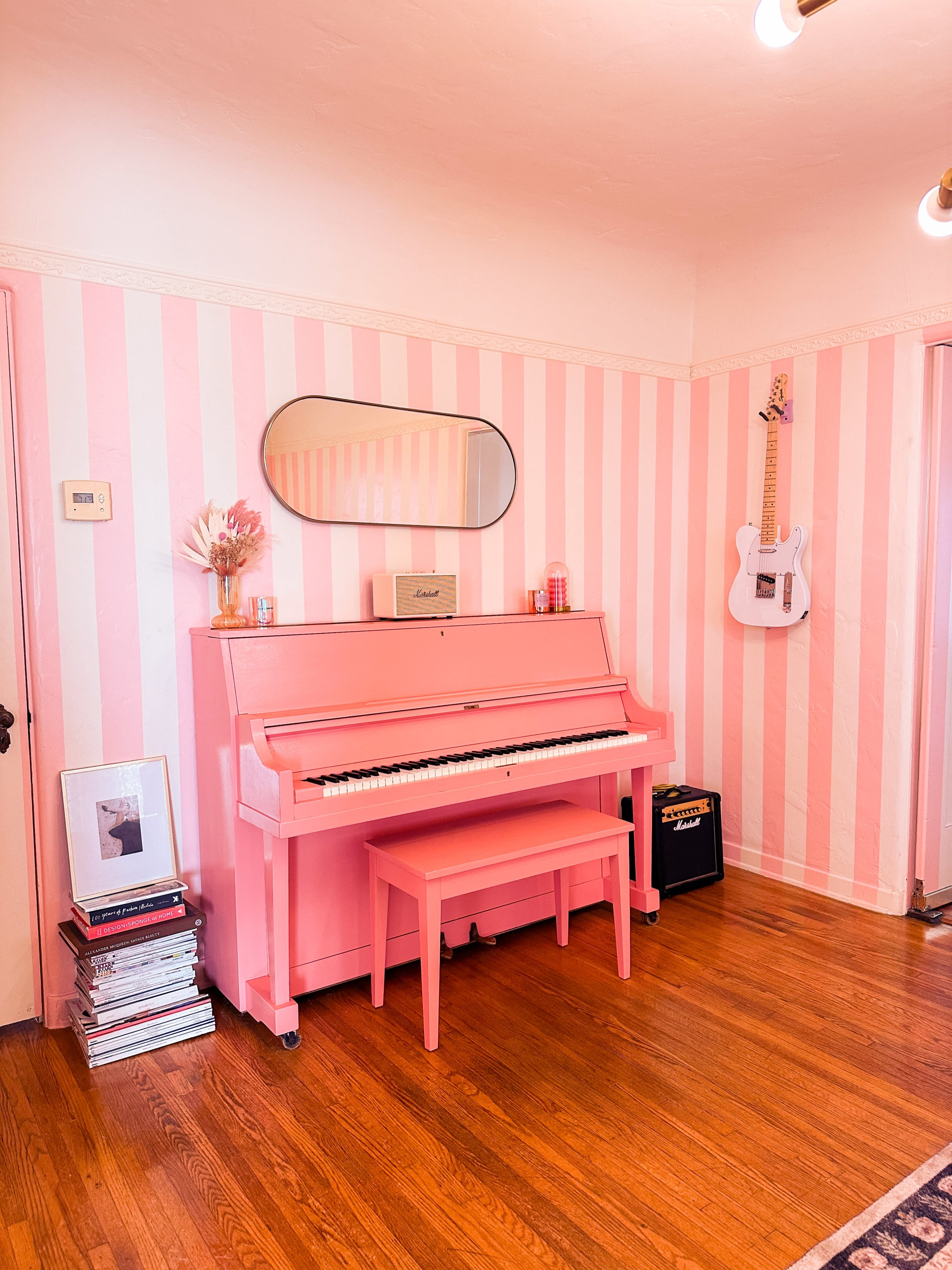 A pink upright piano and matching bench are positioned against striped pink and white walls, with a mirror and a white guitar hung above.