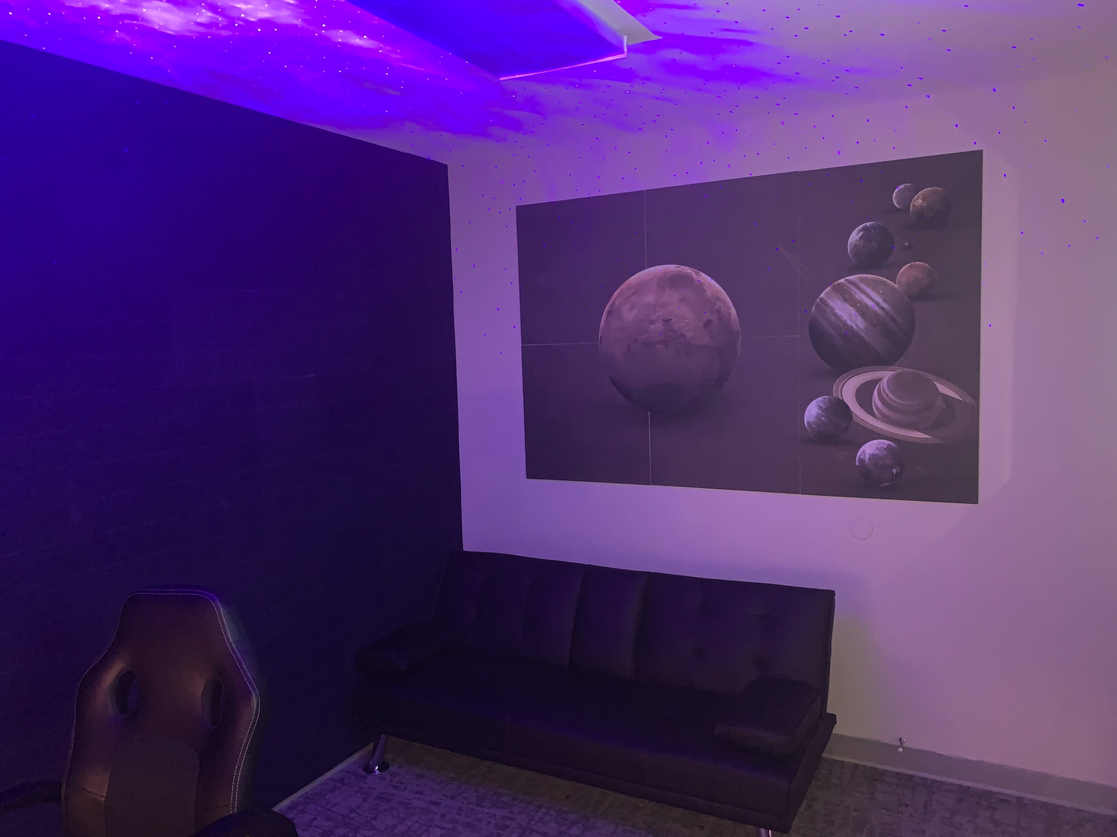 A room with a dark wall, a black couch, and a large mural of planets on the opposite wall, illuminated by purple lighting.