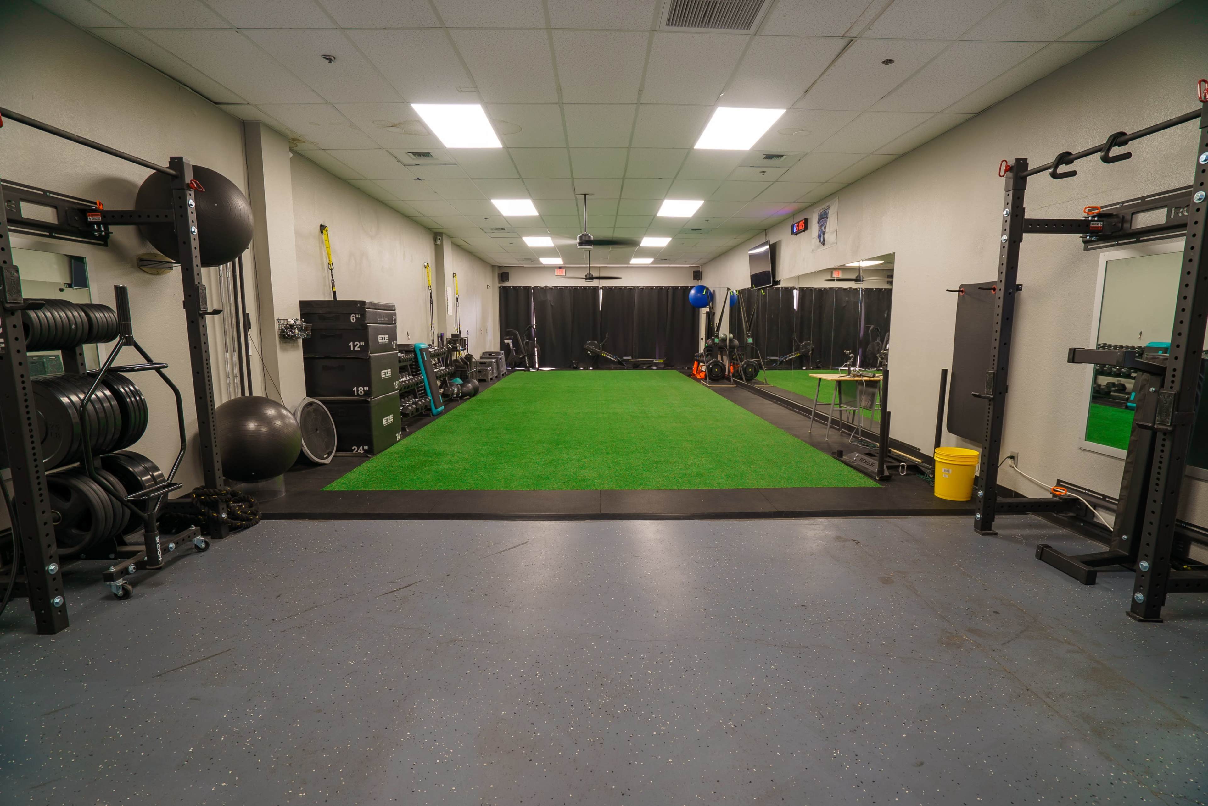 A well-equipped training room with artificial turf, weightlifting equipment along the walls, and a space for various fitness activities.