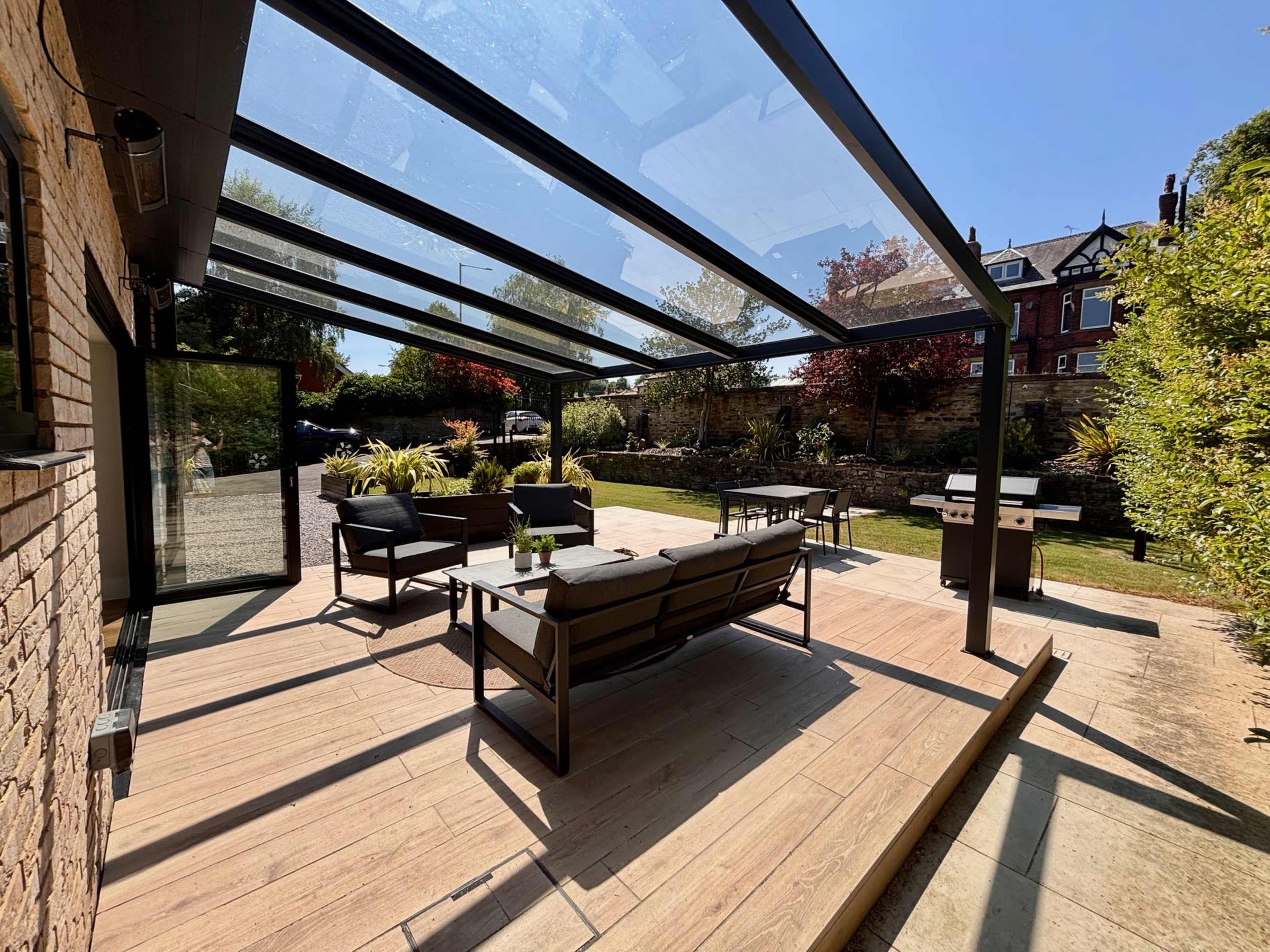 The image shows a modern patio area with a glass-roofed shelter, furnished with seating and a table, overlooking a well-manicured garden.
