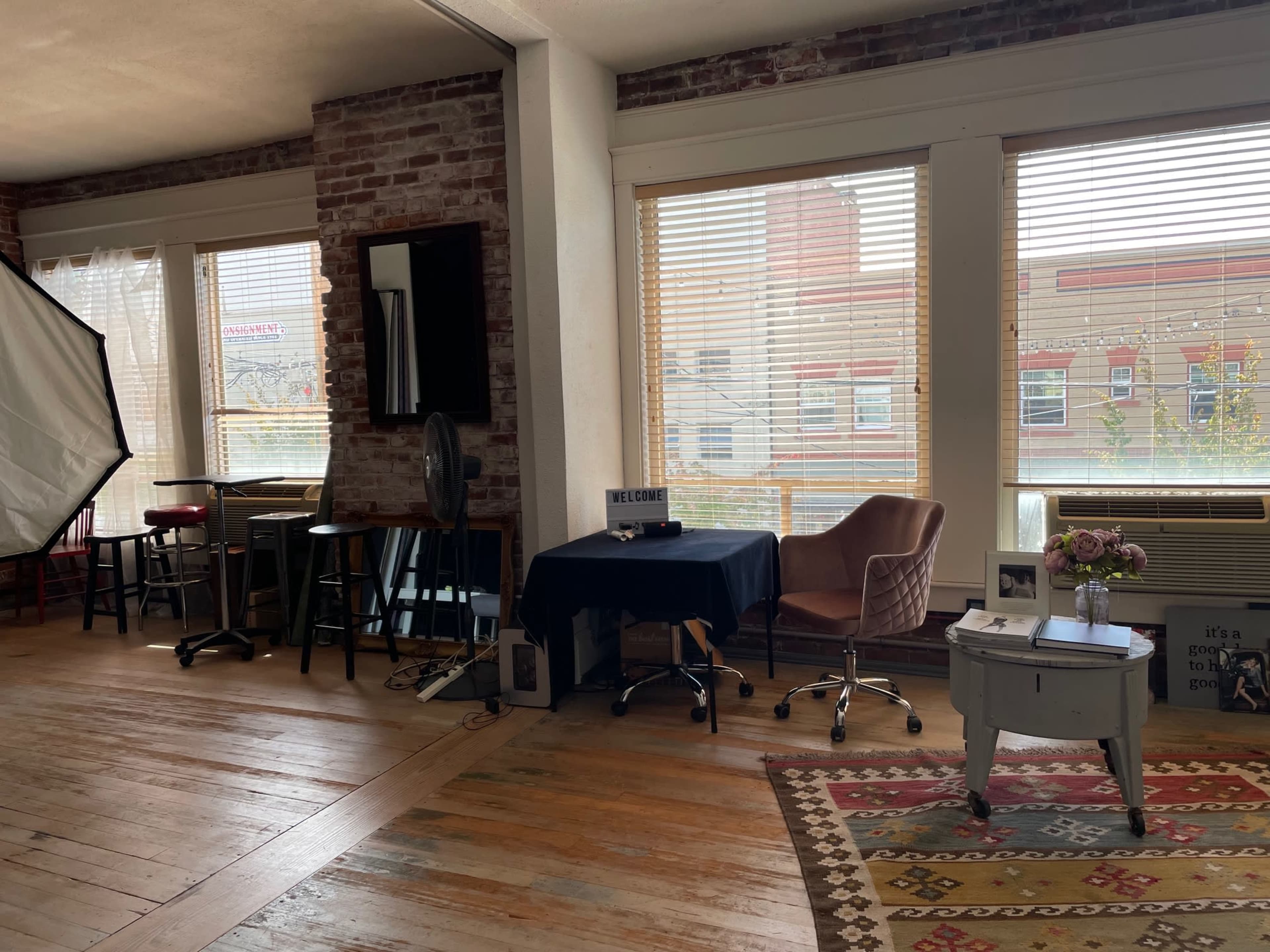 A spacious room features large windows with blinds, hardwood floors, and a mix of furniture including a desk, chairs, and a small table with flowers.