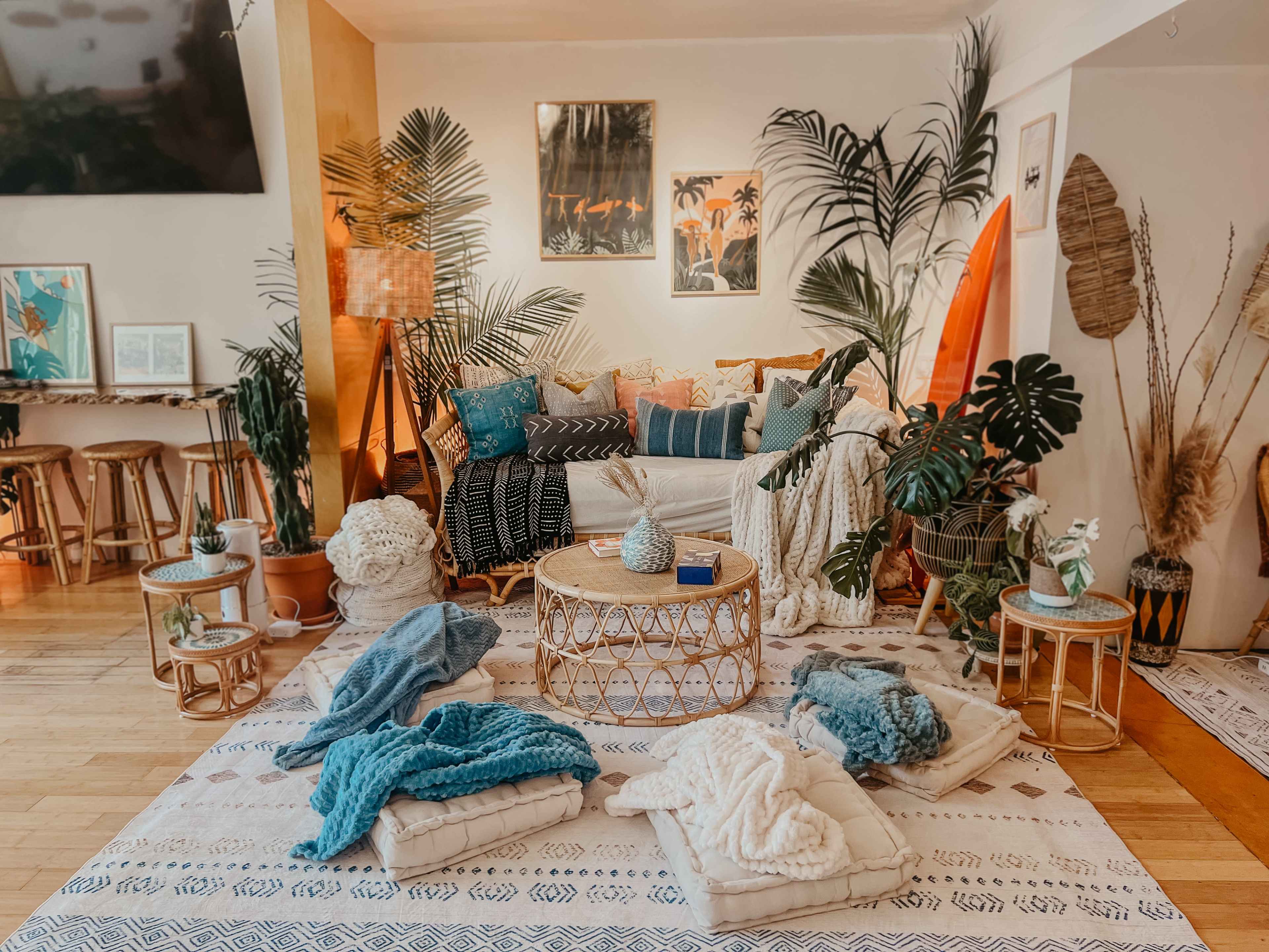 The image features a cozy living space with a sofa adorned with cushions, a round woven coffee table, multiple throws on the floor, and various plants and decor elements.