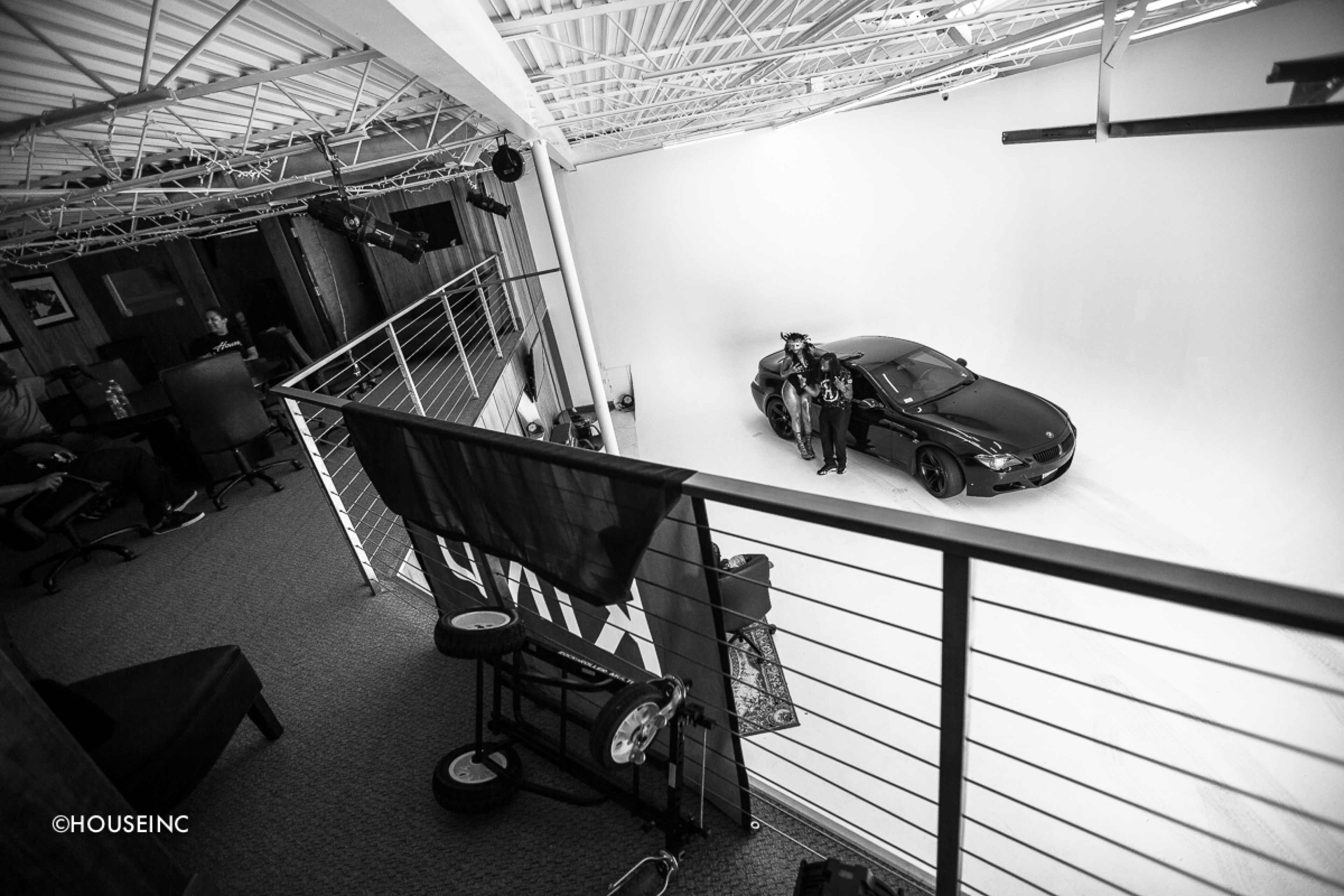 A black car is positioned in a minimalistic studio with white walls while two individuals stand beside it on a raised platform.