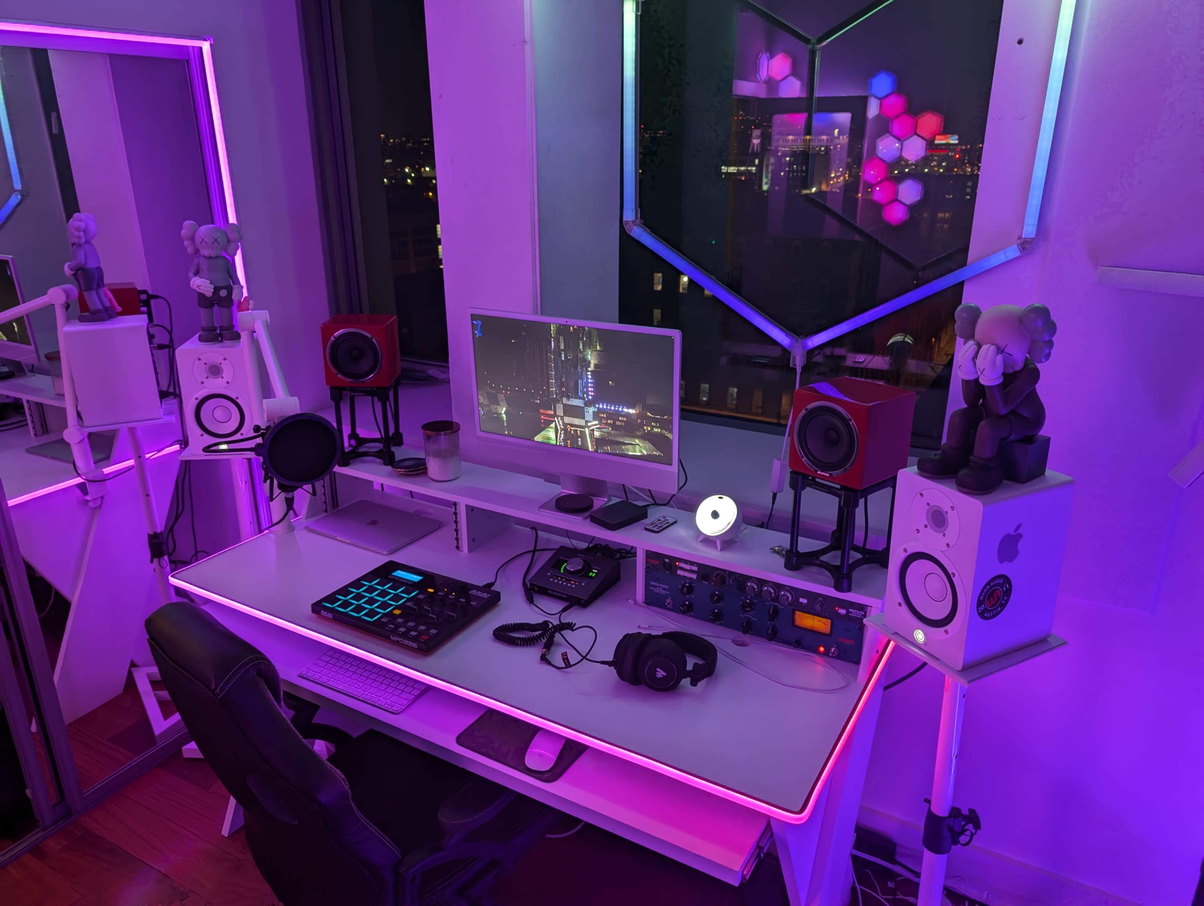 A modern home office setup features a computer monitor, speakers, headphones, and decorative lighting, positioned near a window with a city view.