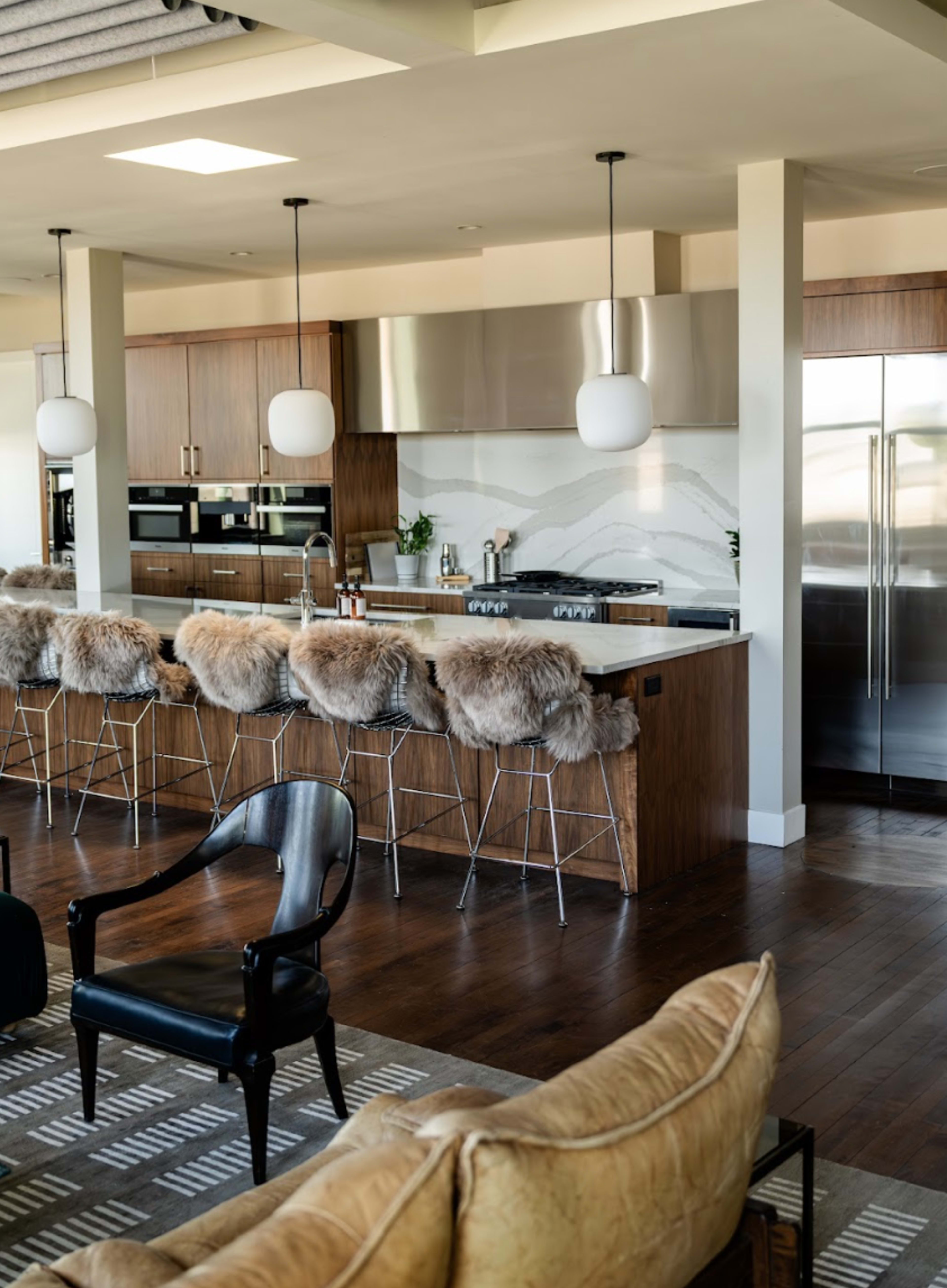 A modern kitchen with a large island bar featuring fur-covered stools, stainless steel appliances, and wooden cabinetry.