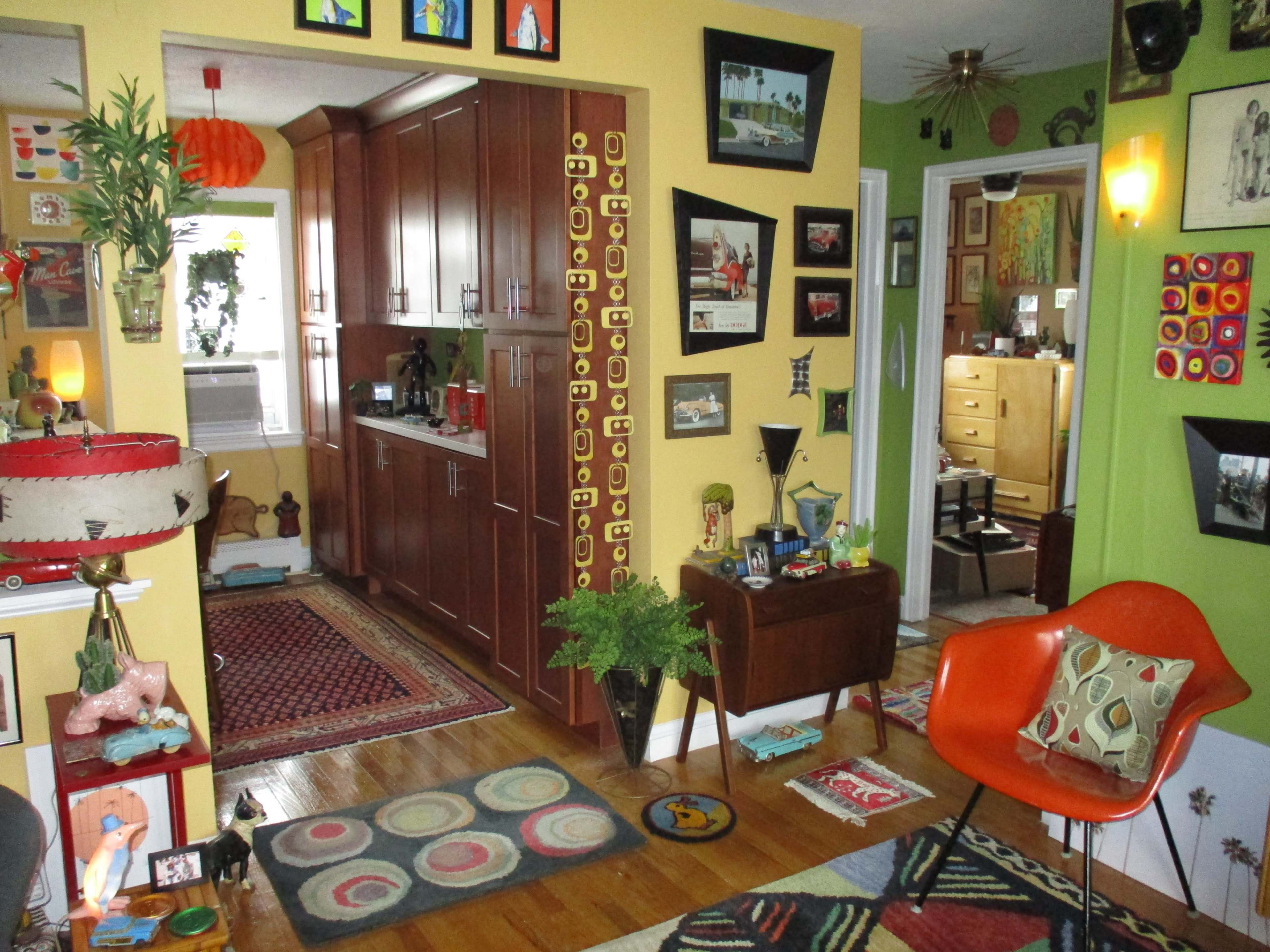 A brightly colored living space featuring a kitchen area, various framed pictures on the walls, a mid-century modern chair, and decorative rugs on wooden flooring.