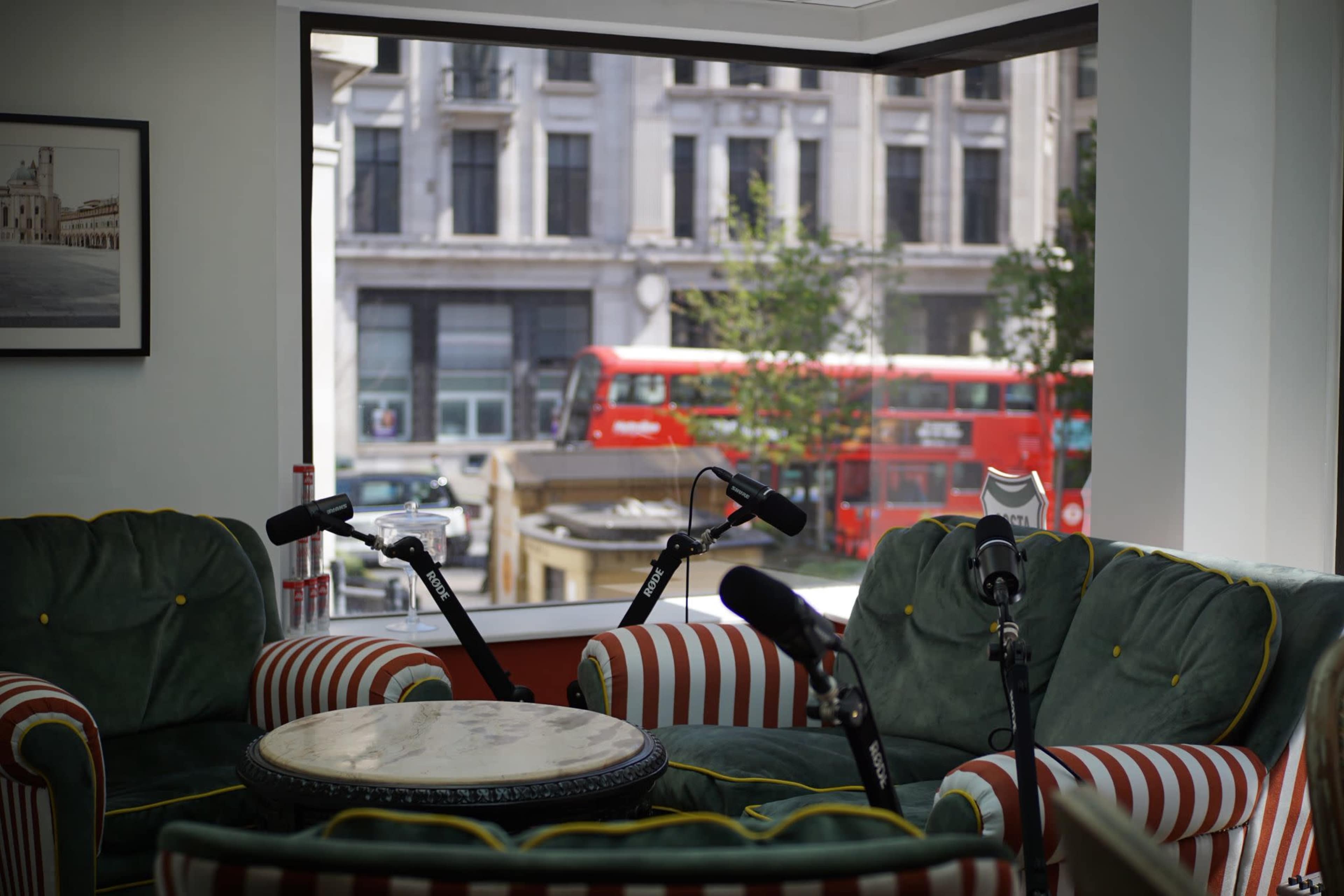 The image shows a cozy seating area with two microphones placed on a green and striped couch, overlooking a street with a red double-decker bus in the background through a large window.